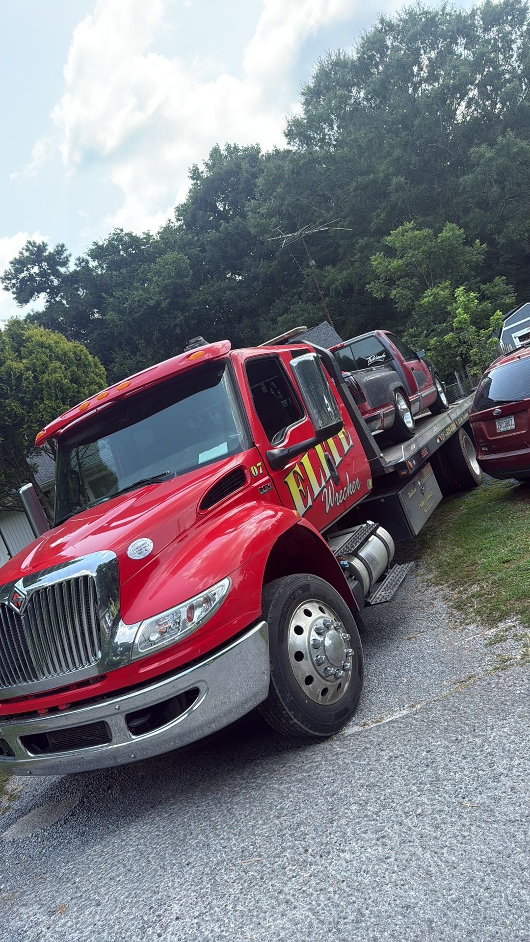Red tow trucks lined up on a gravel lot beside trees and a parked car
