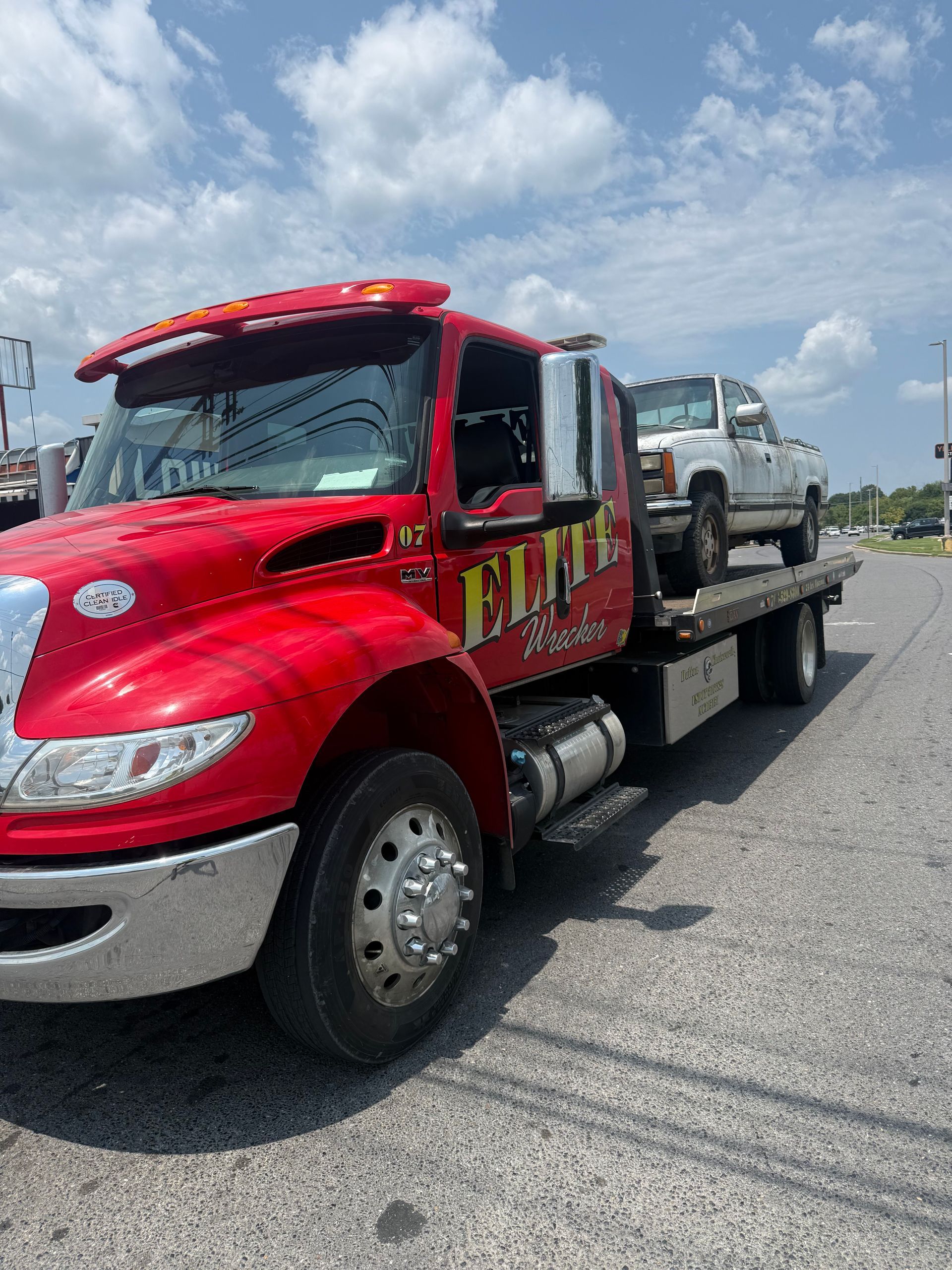 Red flatbed tow truck carrying a white SUV on a highway under a partly cloudy sky