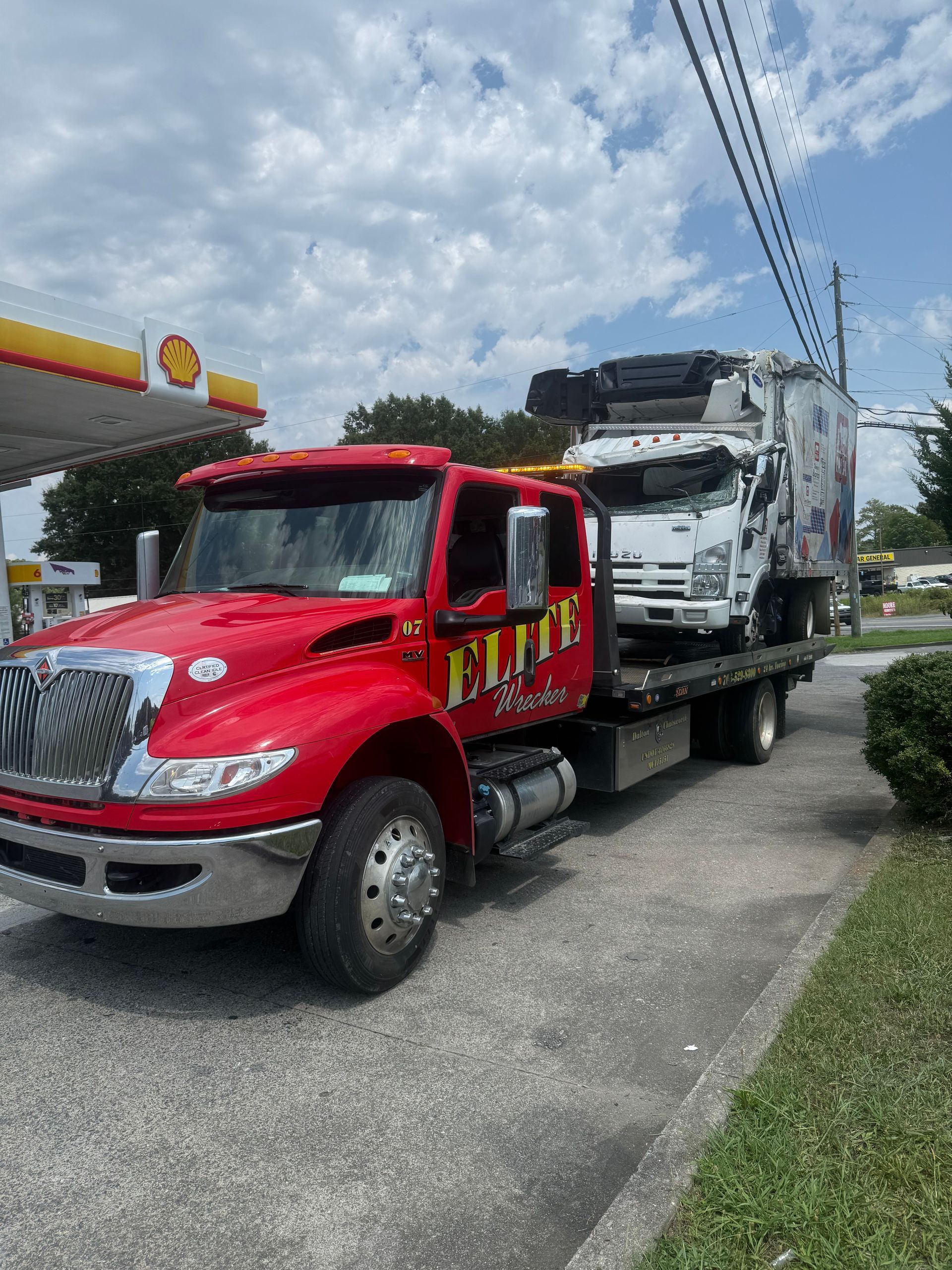 Red tow truck hauling a white semi-truck on a roadside under a cloudy sky.