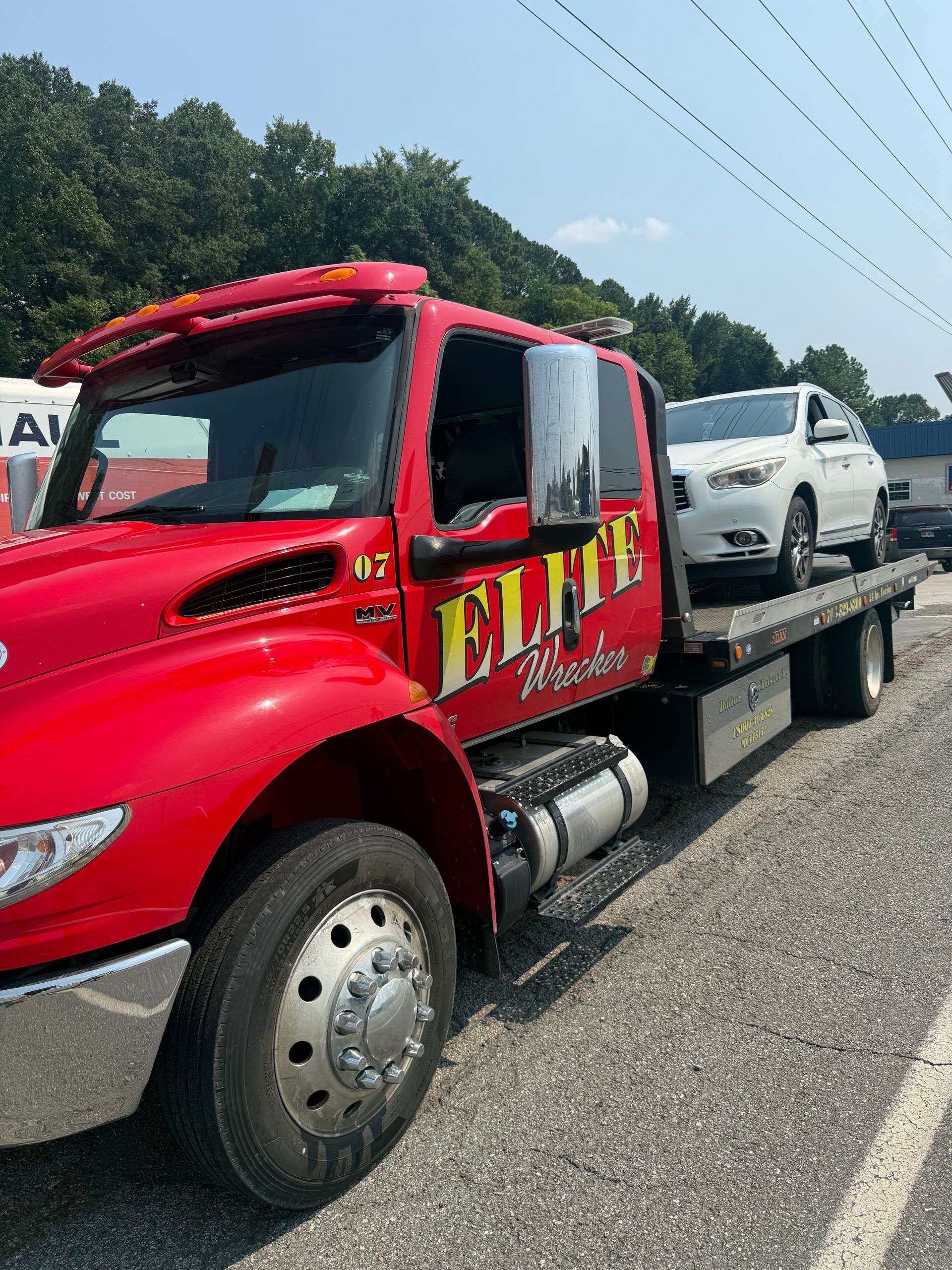 Red tow truck hauling a white pickup on a flatbed along a highway, with trees and power lines in the background