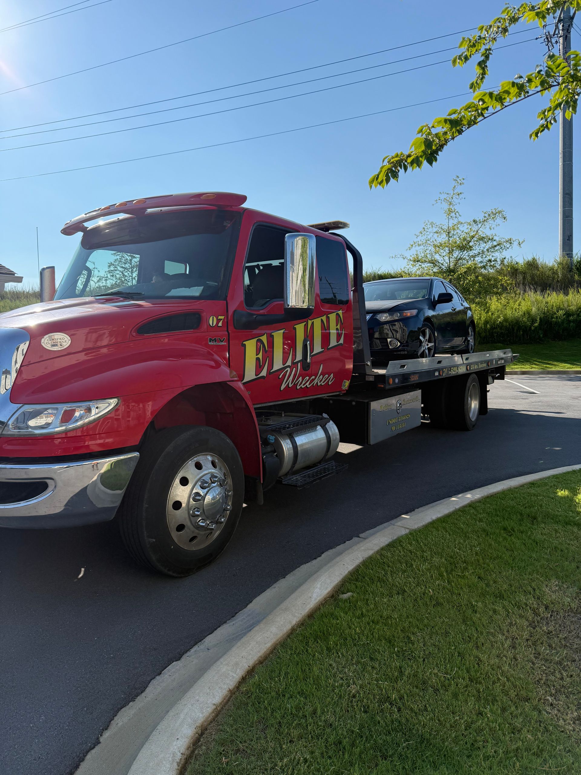 Red tow truck with a damaged black car on its flatbed, parked by a curved road on a sunny day