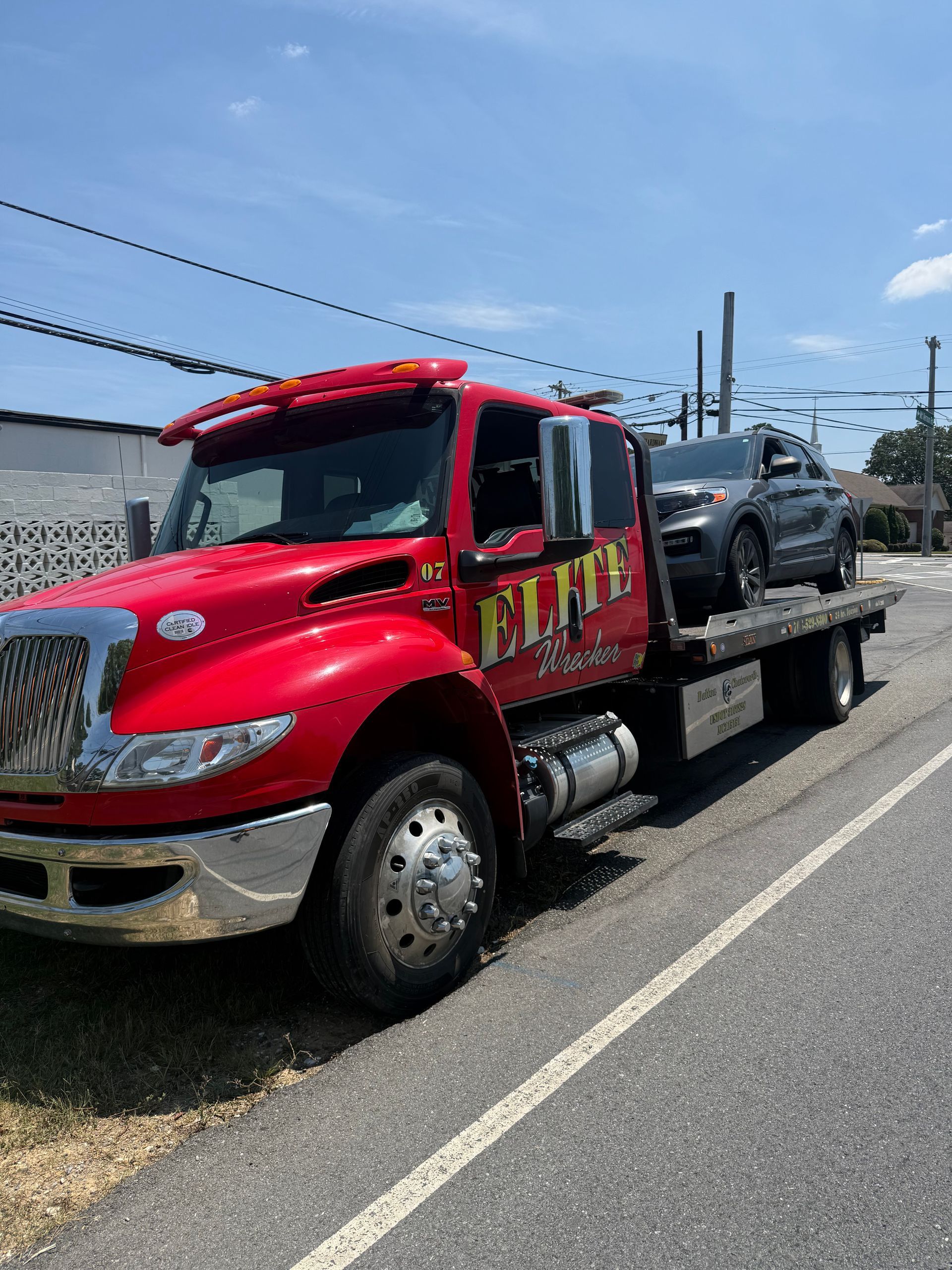 Red tow truck carrying a dark SUV on a flatbed along a roadside under a blue sky.