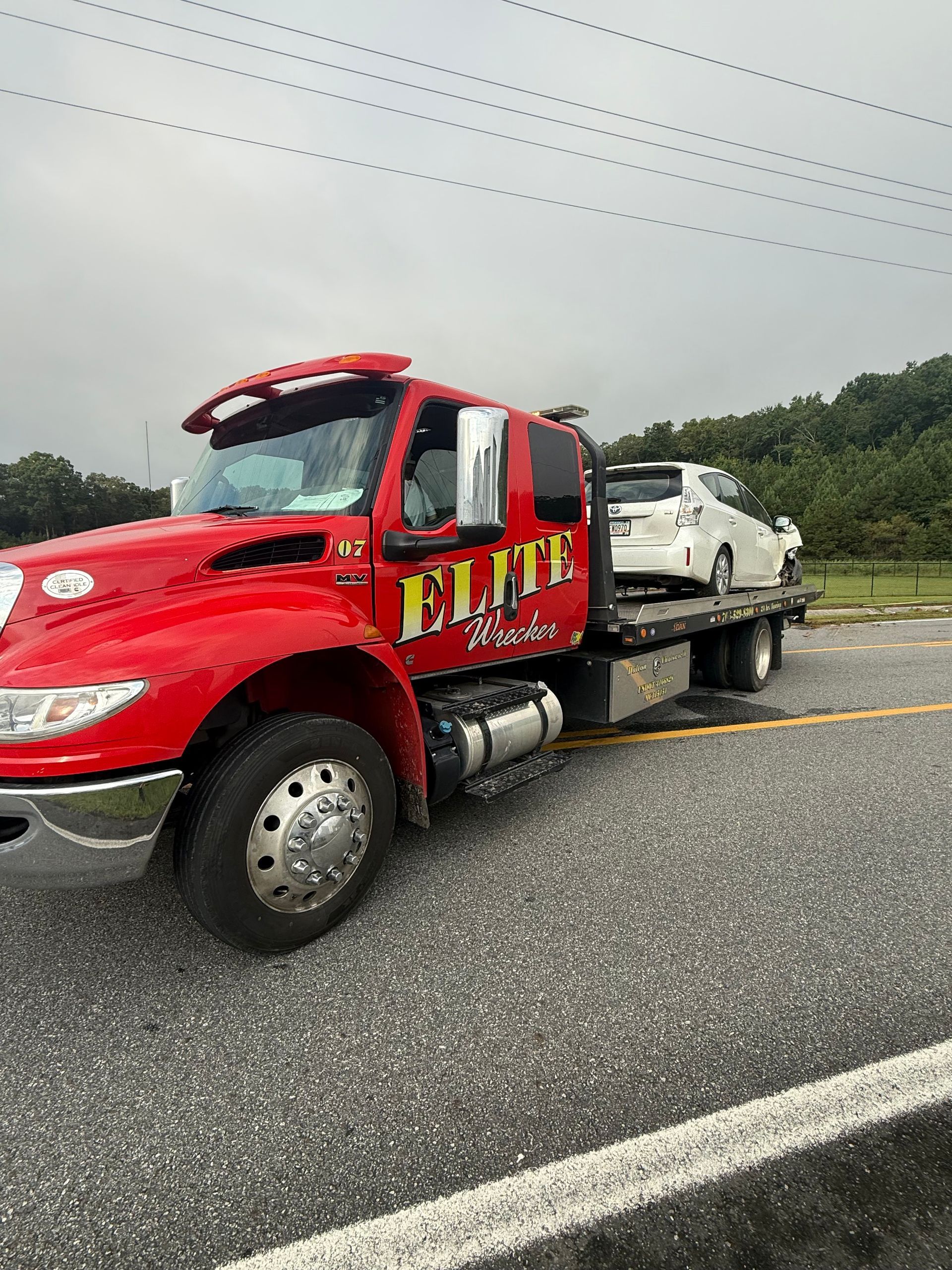 Red tow truck carrying a damaged white car on a wet highway shoulder under cloudy skies