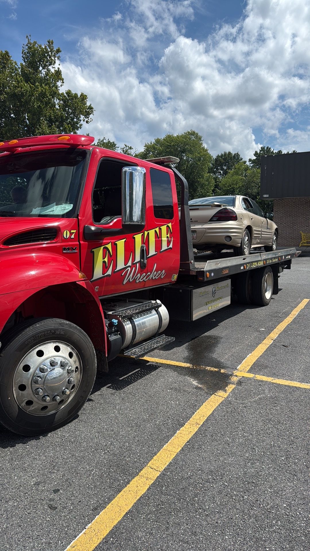 Red tow truck hauling a beige car in a parking lot under a cloudy sky.
