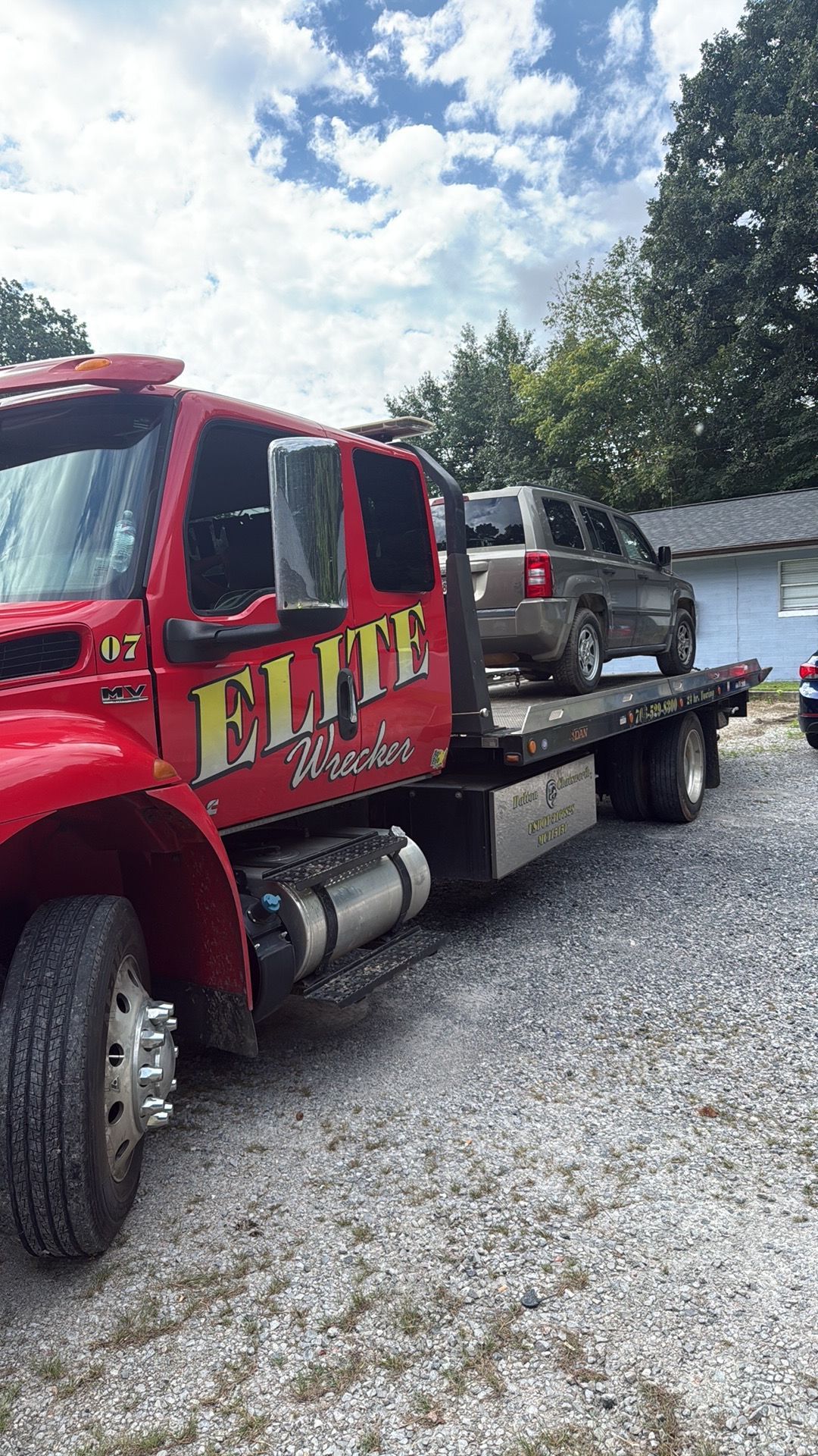 Red tow truck carrying a dark SUV on a flatbed beside a white house on a gravel driveway