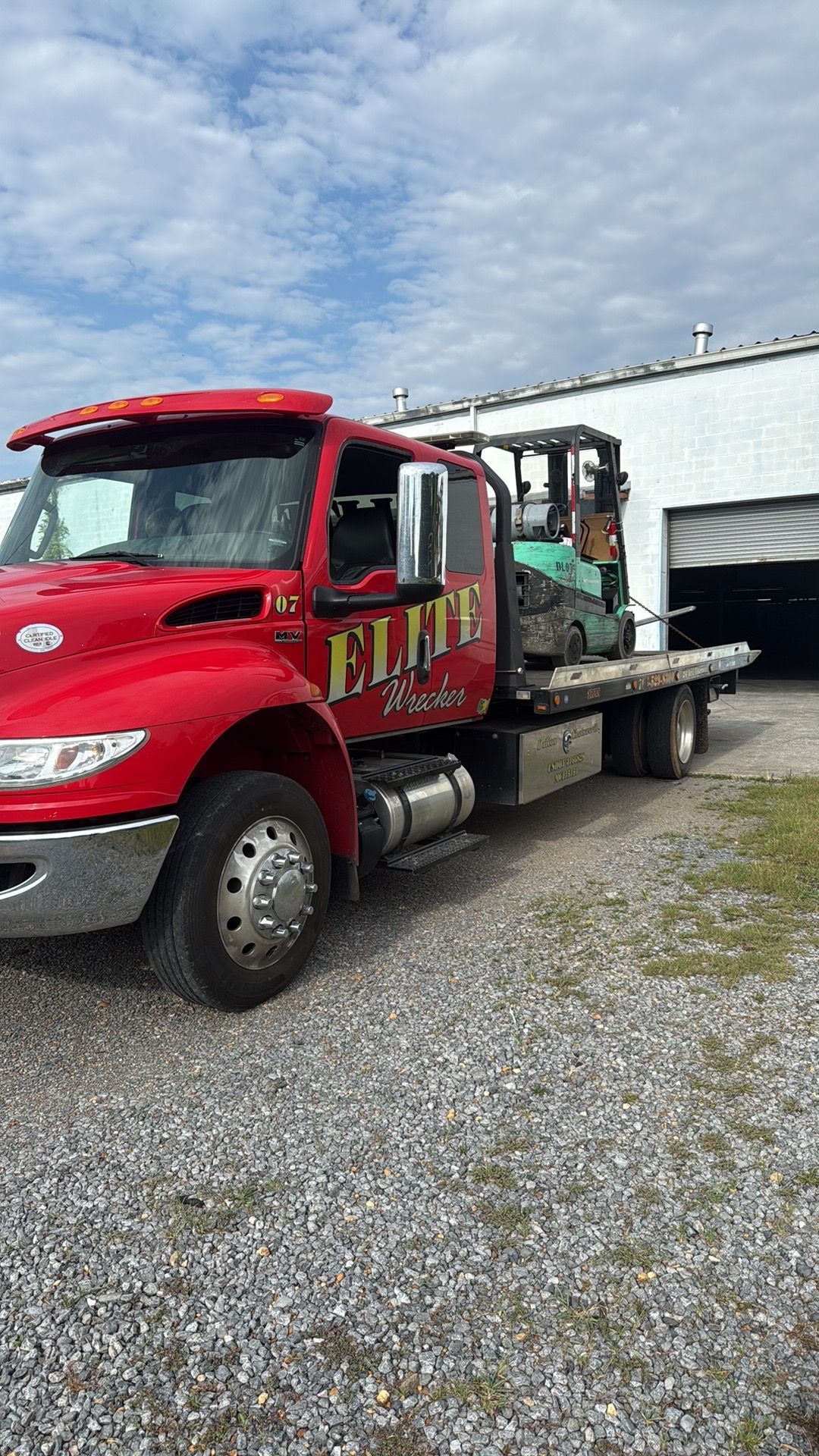 Red flatbed tow truck parked on gravel beside a warehouse, carrying equipment.