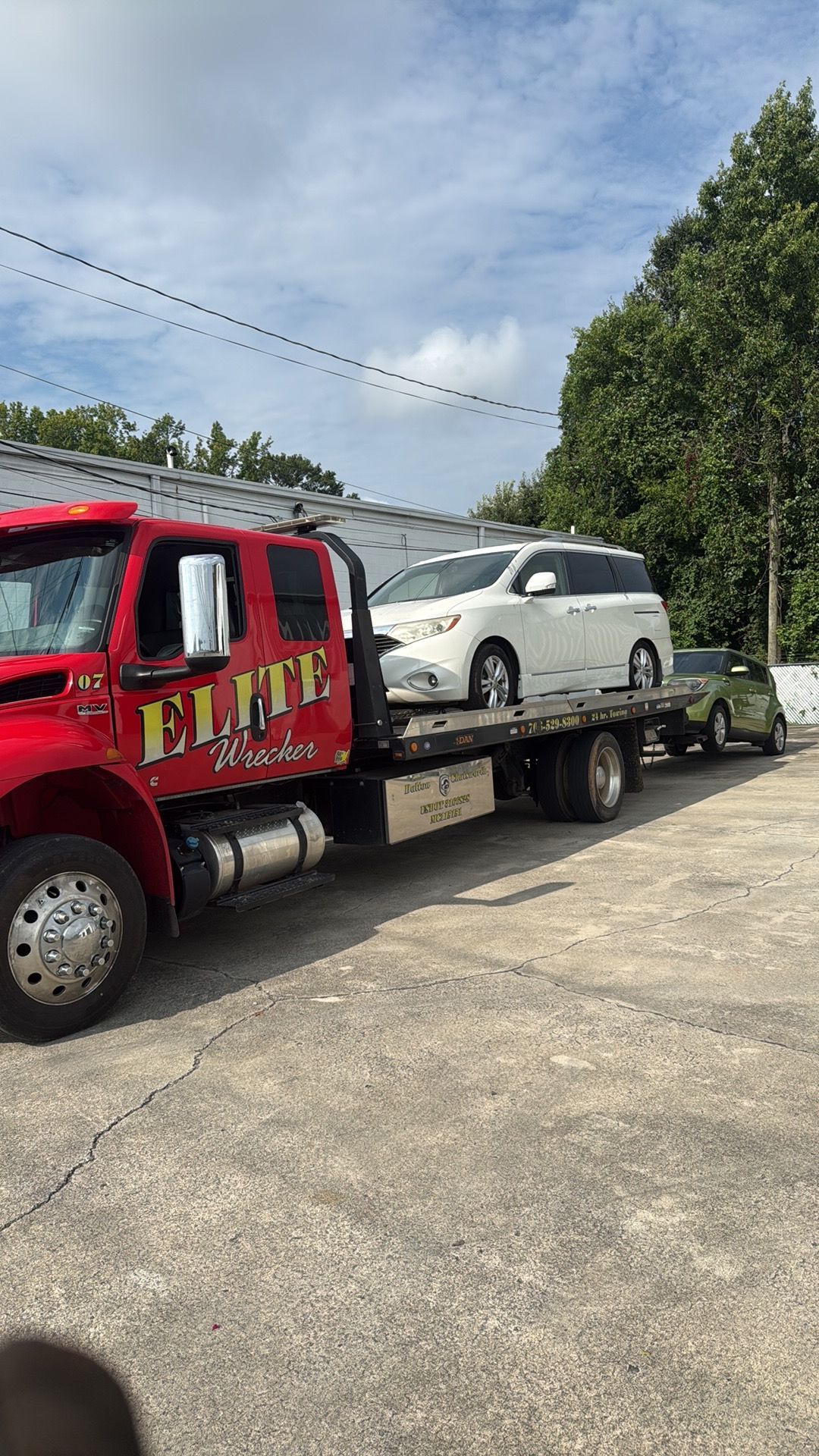Red tow truck hauling a white SUV on a flatbed in a gravel lot under a cloudy sky