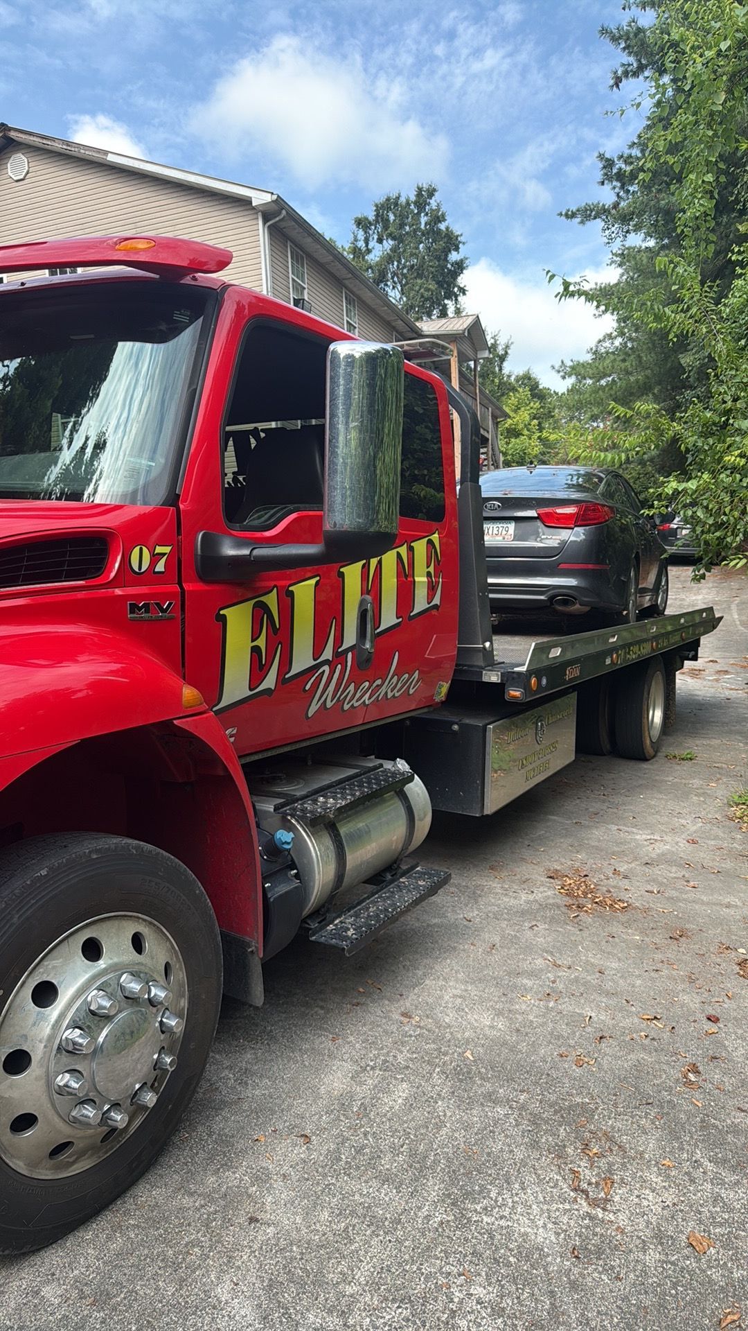 Red tow truck hauling a black SUV on a flatbed beside a building and trees
