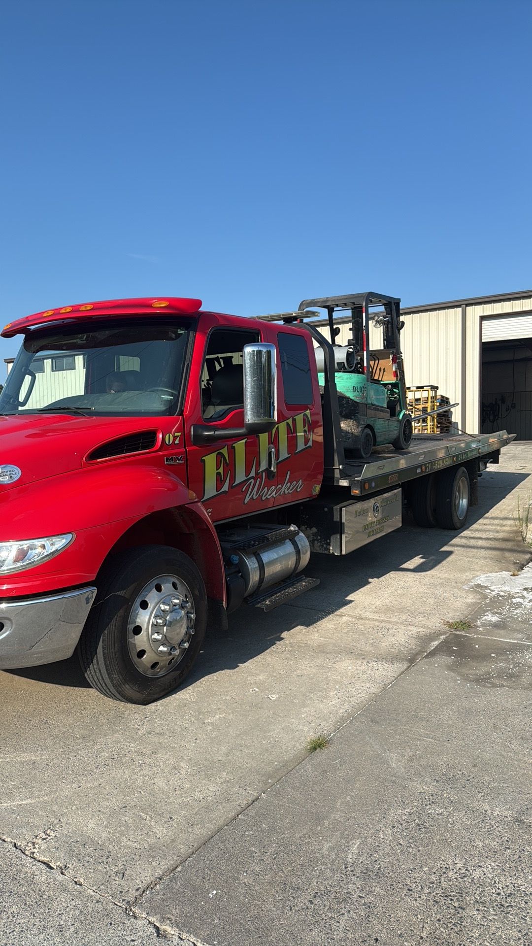 Red flatbed tow truck parked outside a garage on a sunny day.