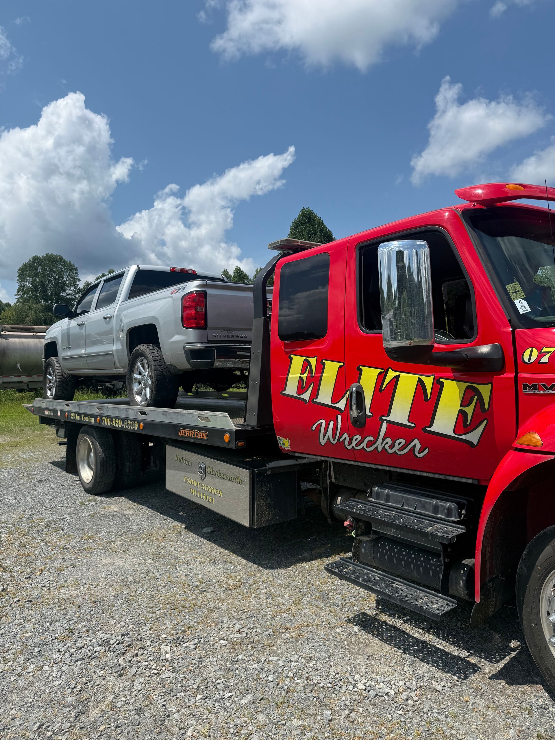 Red tow truck hauling a silver pickup on a flatbed in a gravel lot under a blue sky.