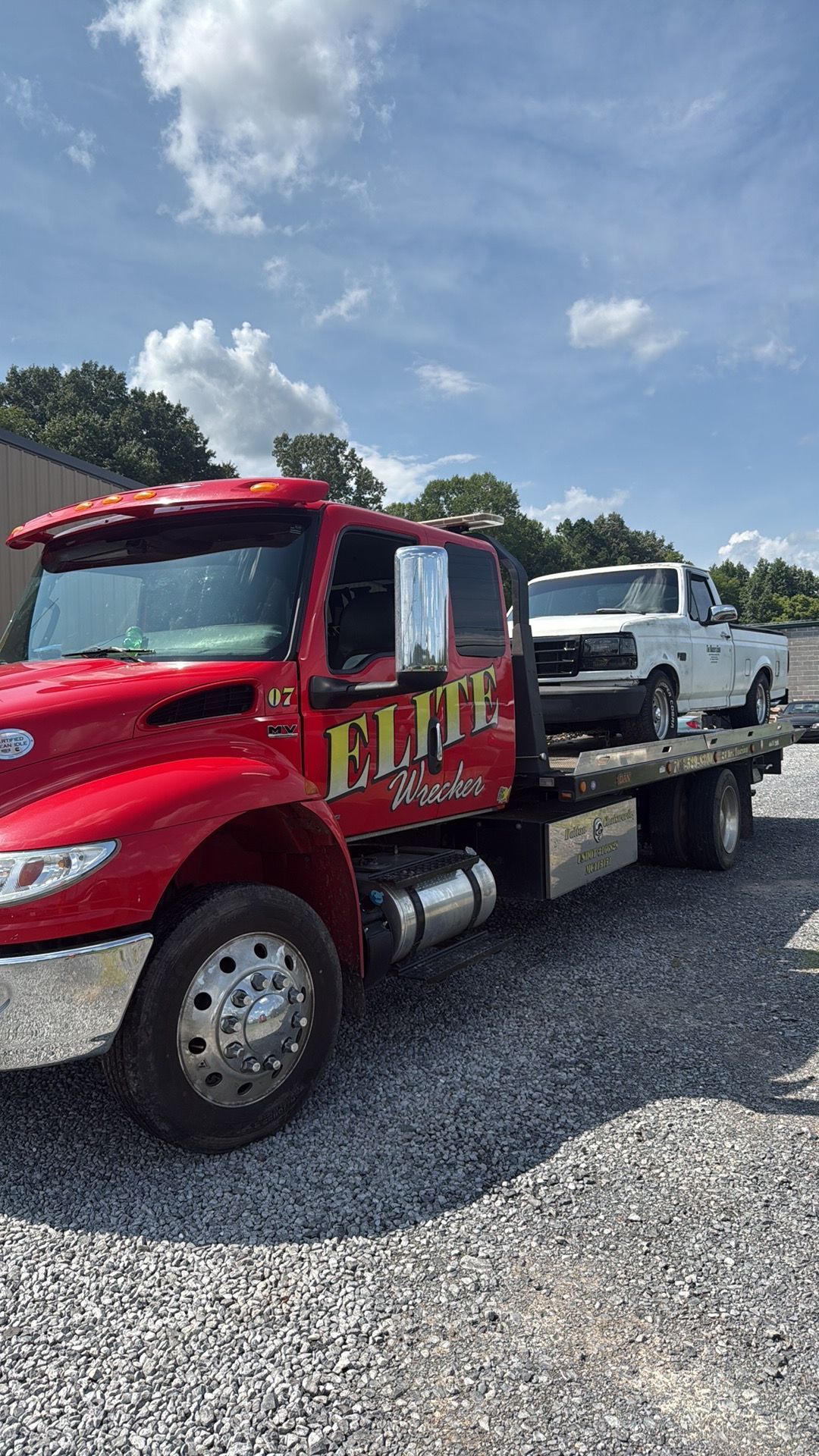 Red flatbed tow truck carrying a white pickup on a gravel lot under a blue sky