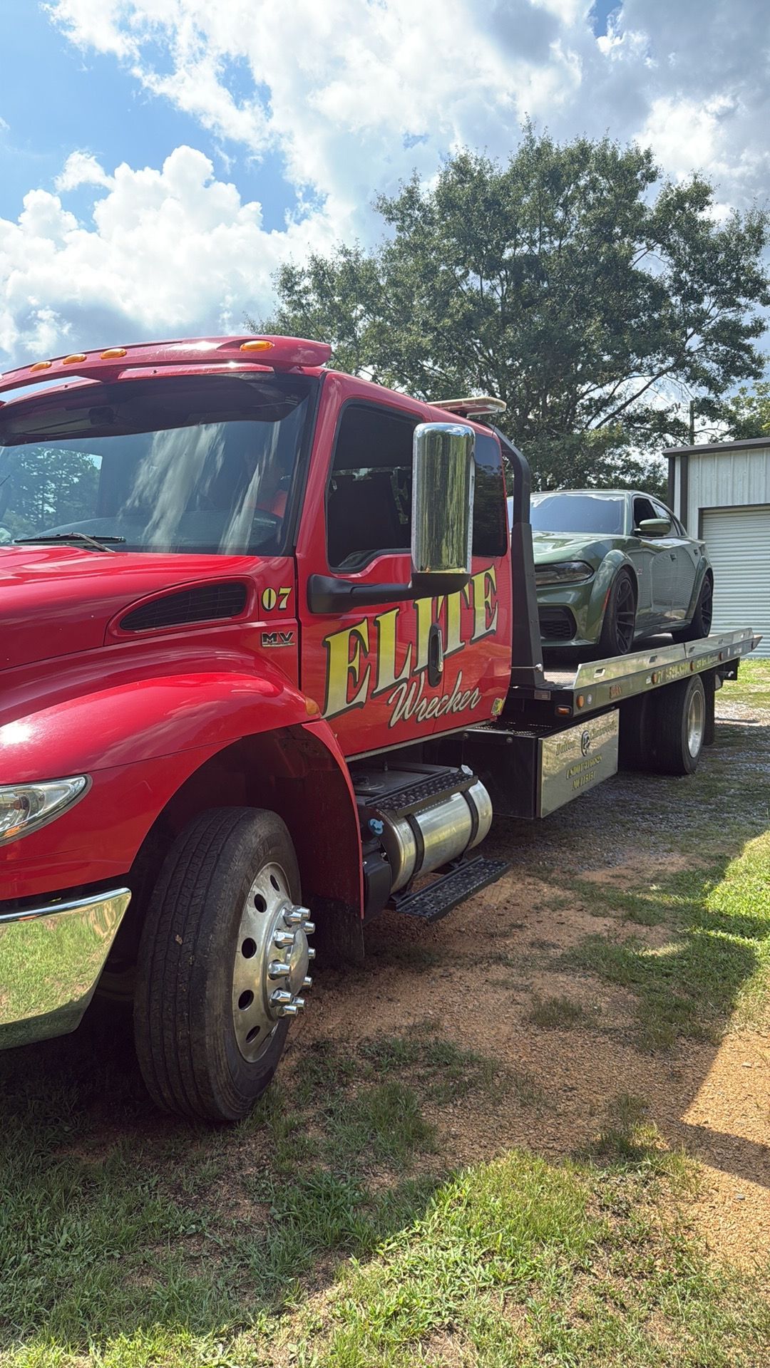 Red flatbed tow truck parked on grass beside a white shed under a cloudy sky