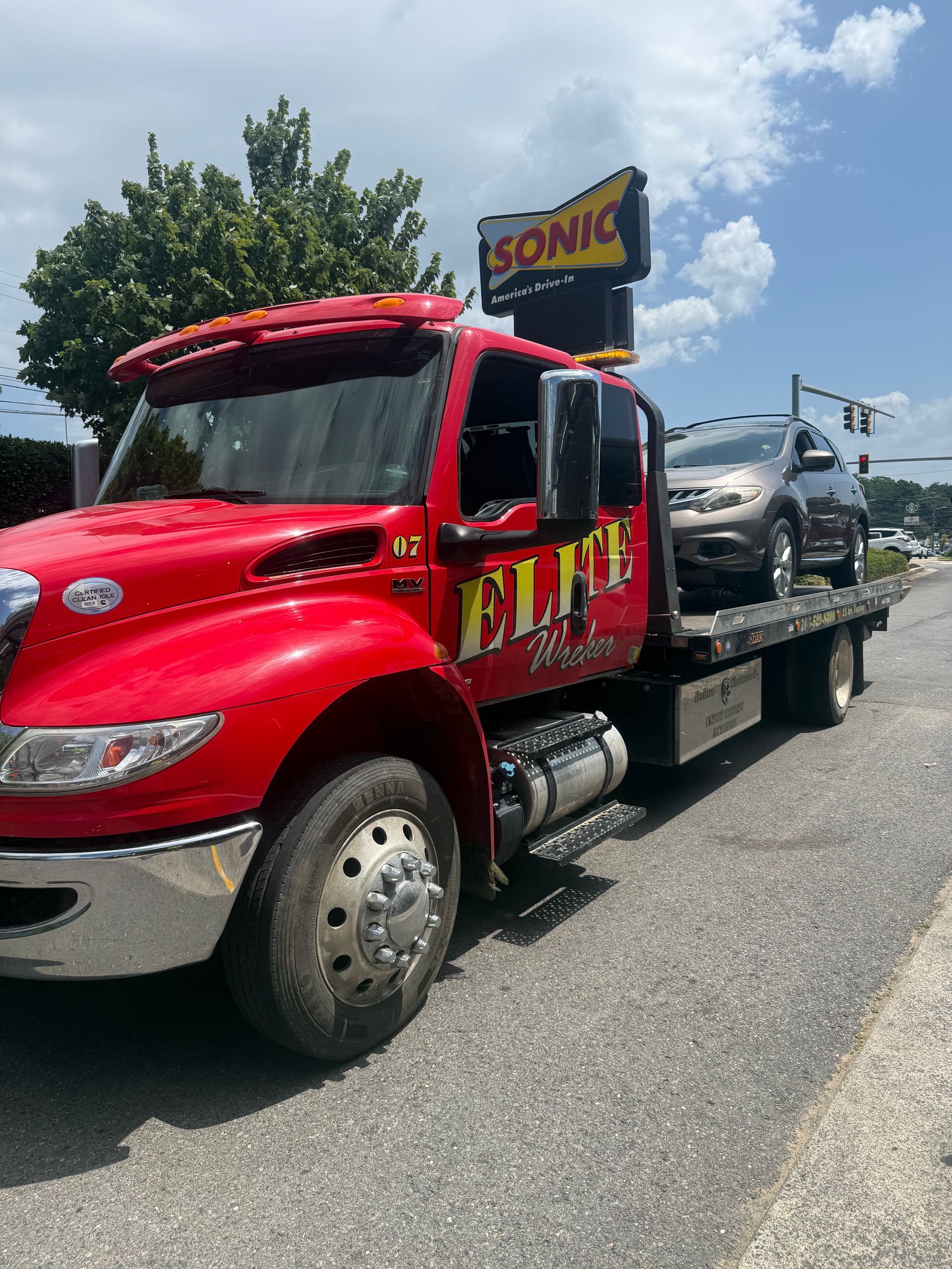 Red flatbed tow truck carrying a black car on a sunny roadside, with a Sonic sign above.