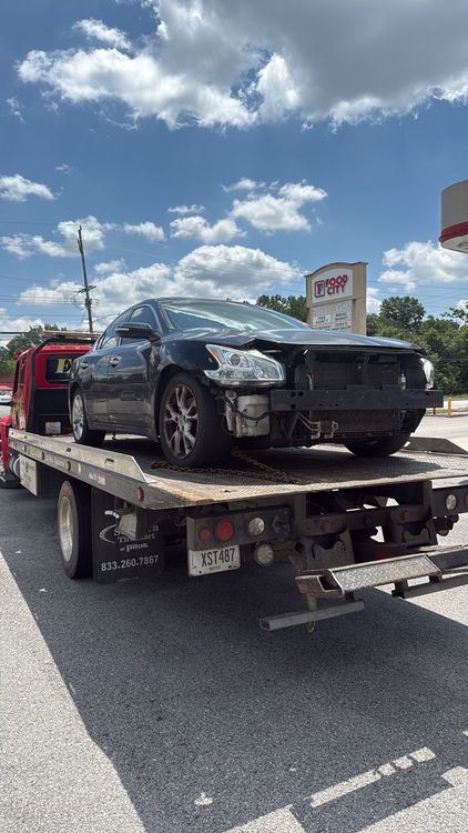 Damaged black car on a tow truck parked by a roadside under a cloudy sky