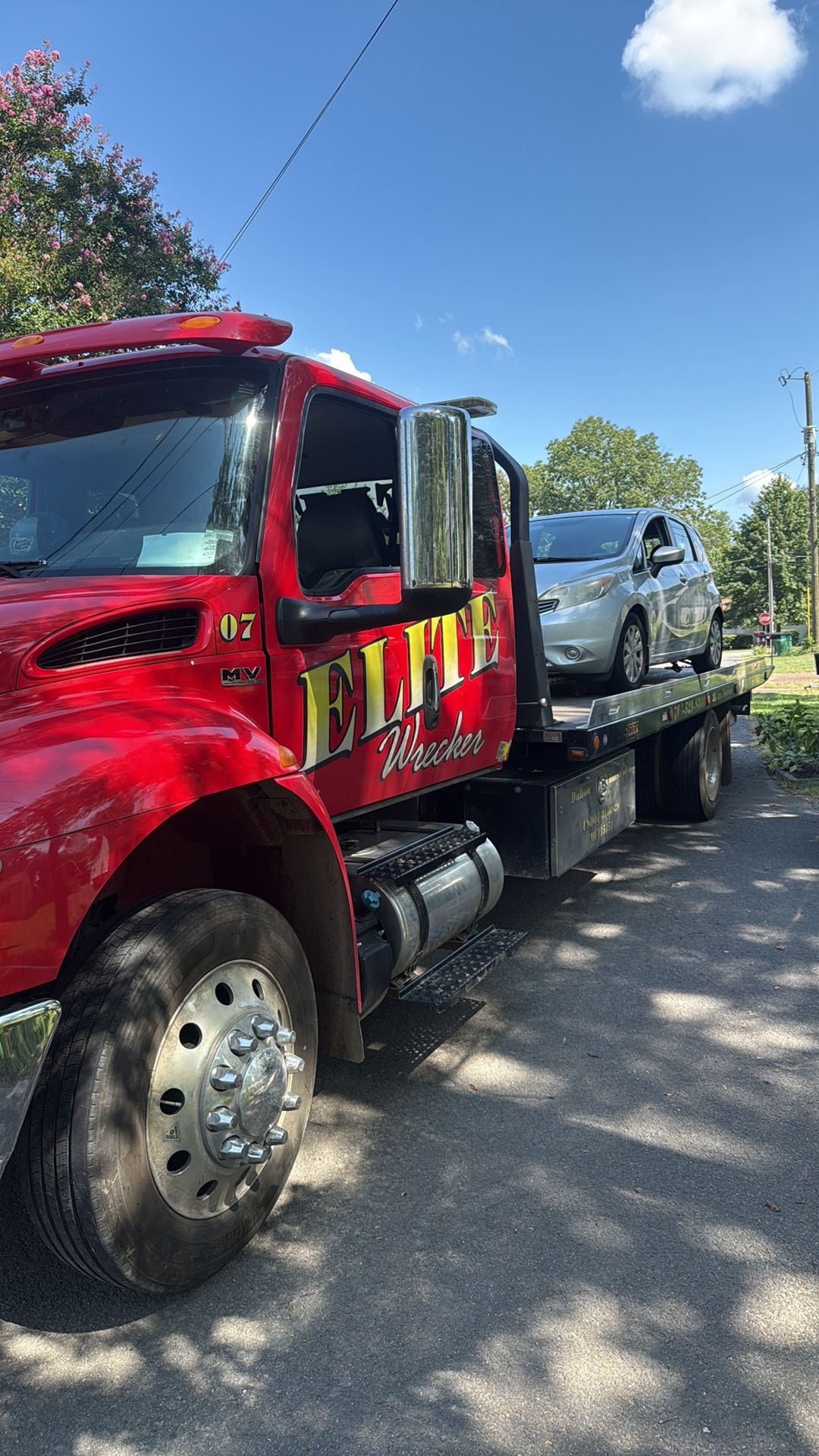 Red tow truck hauling a silver pickup on a flatbed beside a tree-lined road.
