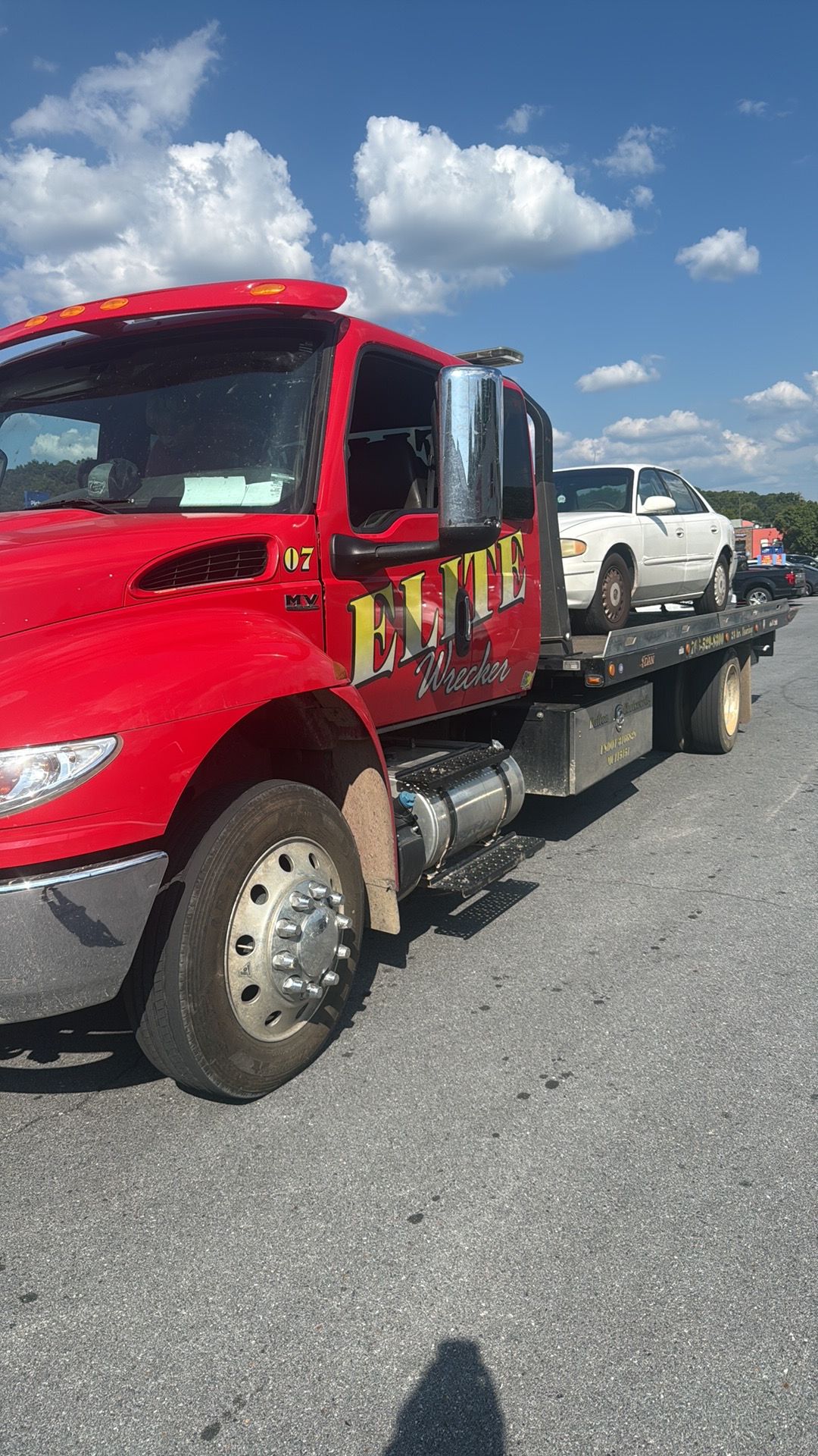 Red tow truck carrying a white car on a gravel lot under a blue sky