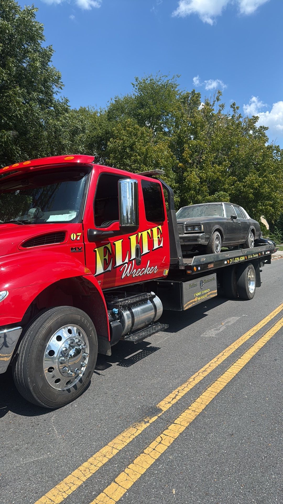 Red tow truck hauling a damaged gray SUV on a road with double yellow lines under a blue sky