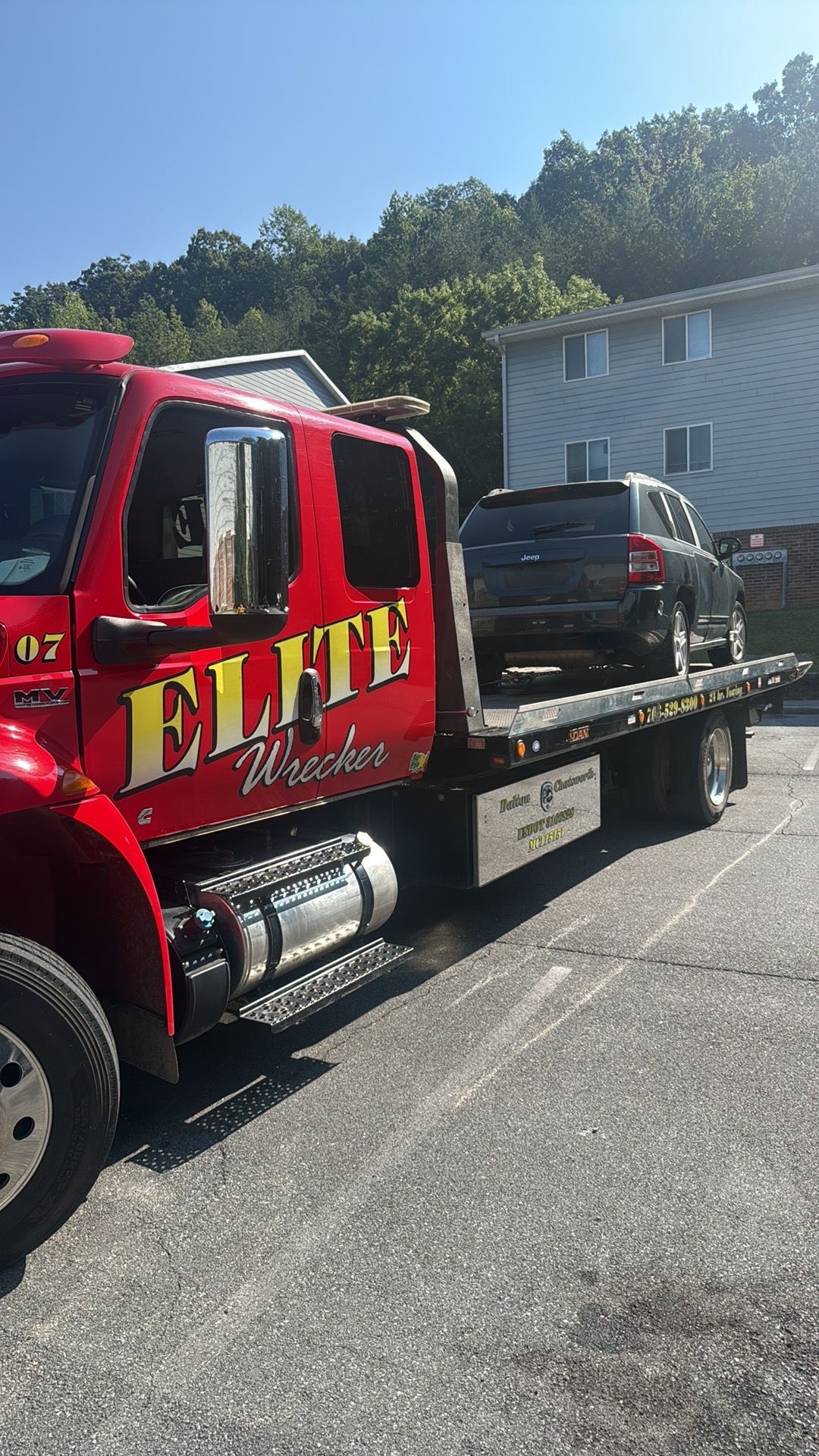 Red flatbed tow truck hauling a black SUV on a sunny roadside near trees and a house