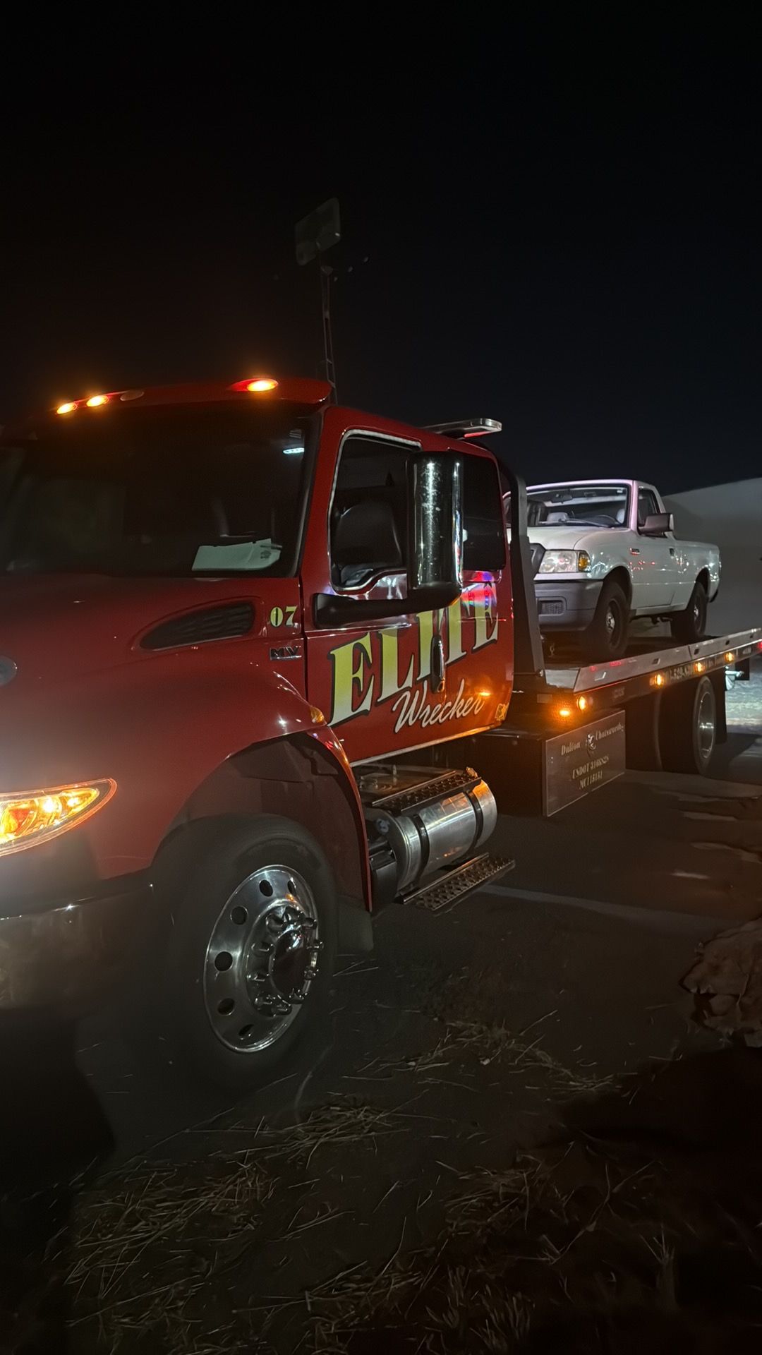 Red tow truck at night hauling a white pickup truck on a flatbed trailer.