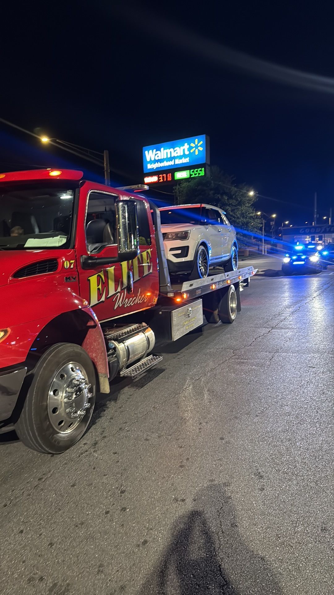 Red tow truck hauling a white car on a wet road at night, with police lights in the distance