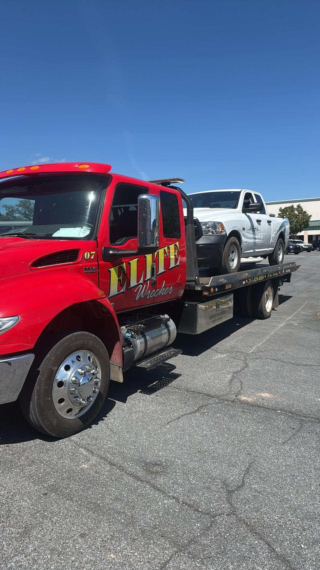 Red tow truck hauling a silver pickup in a parking lot under a clear blue sky