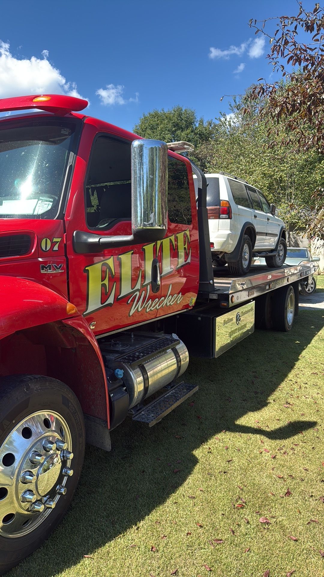 Red tow truck carrying a white SUV on a flatbed in a grassy area under a blue sky
