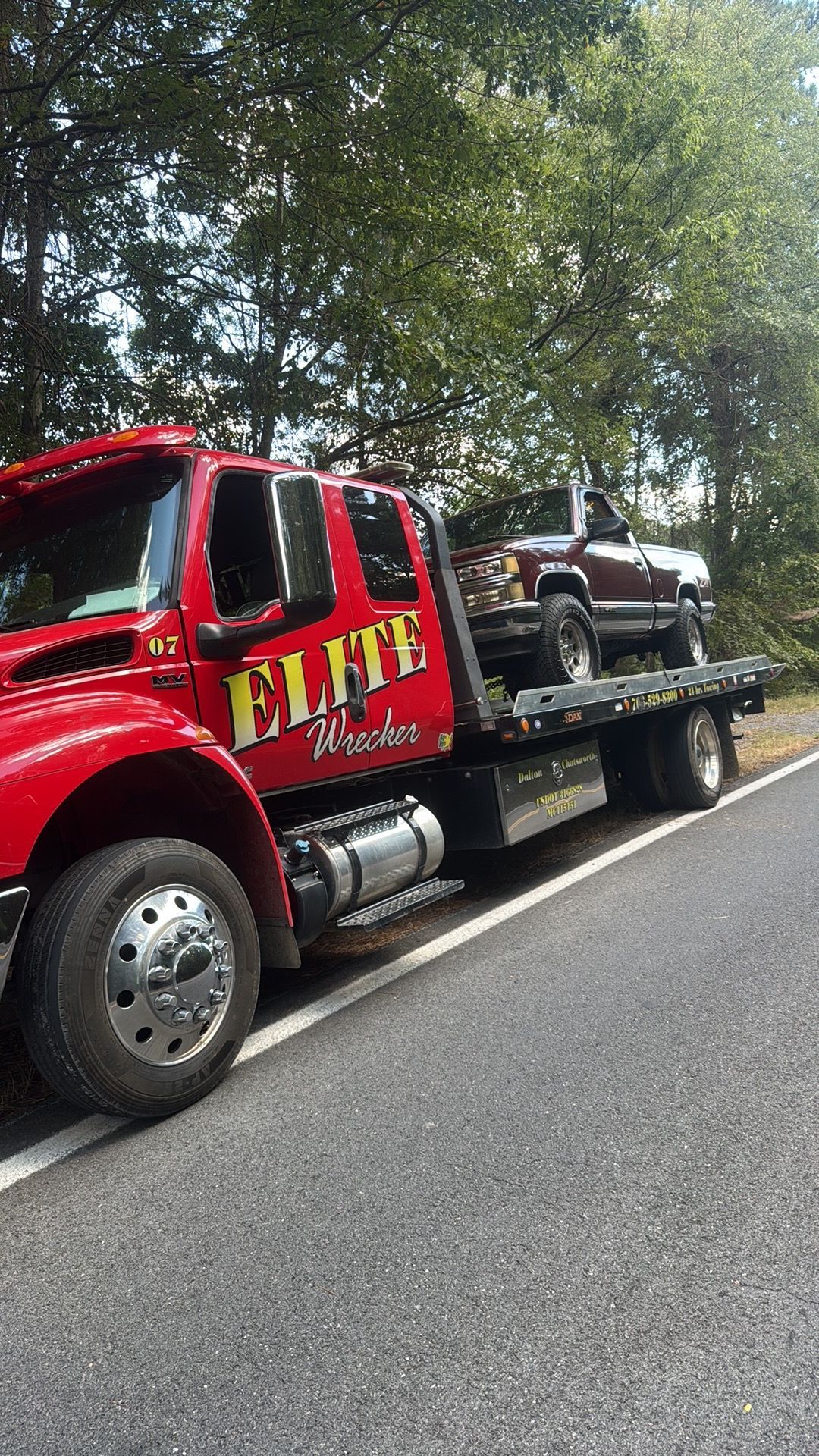 Red tow truck carrying a brown SUV on a flatbed beside a wooded road