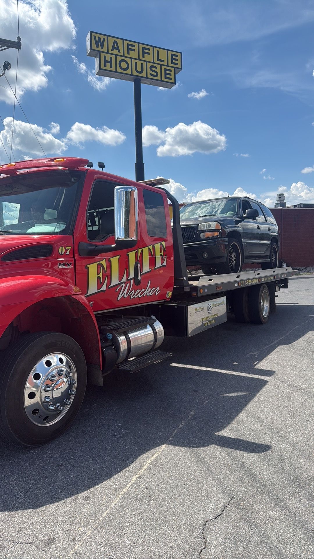 Red tow truck hauling a dark car on a flatbed under a blue sky at a parking lot entrance