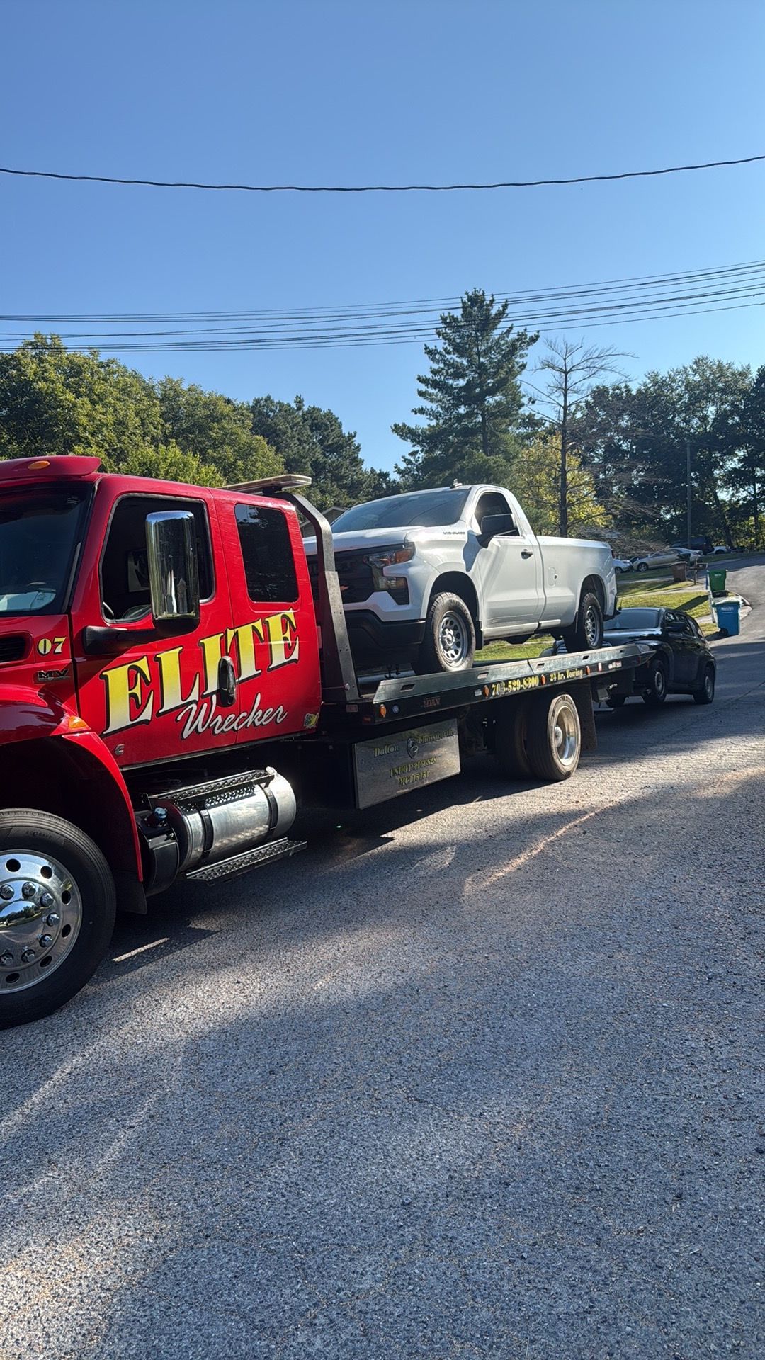 Red flatbed tow truck carrying a white pickup on a sunny suburban street
