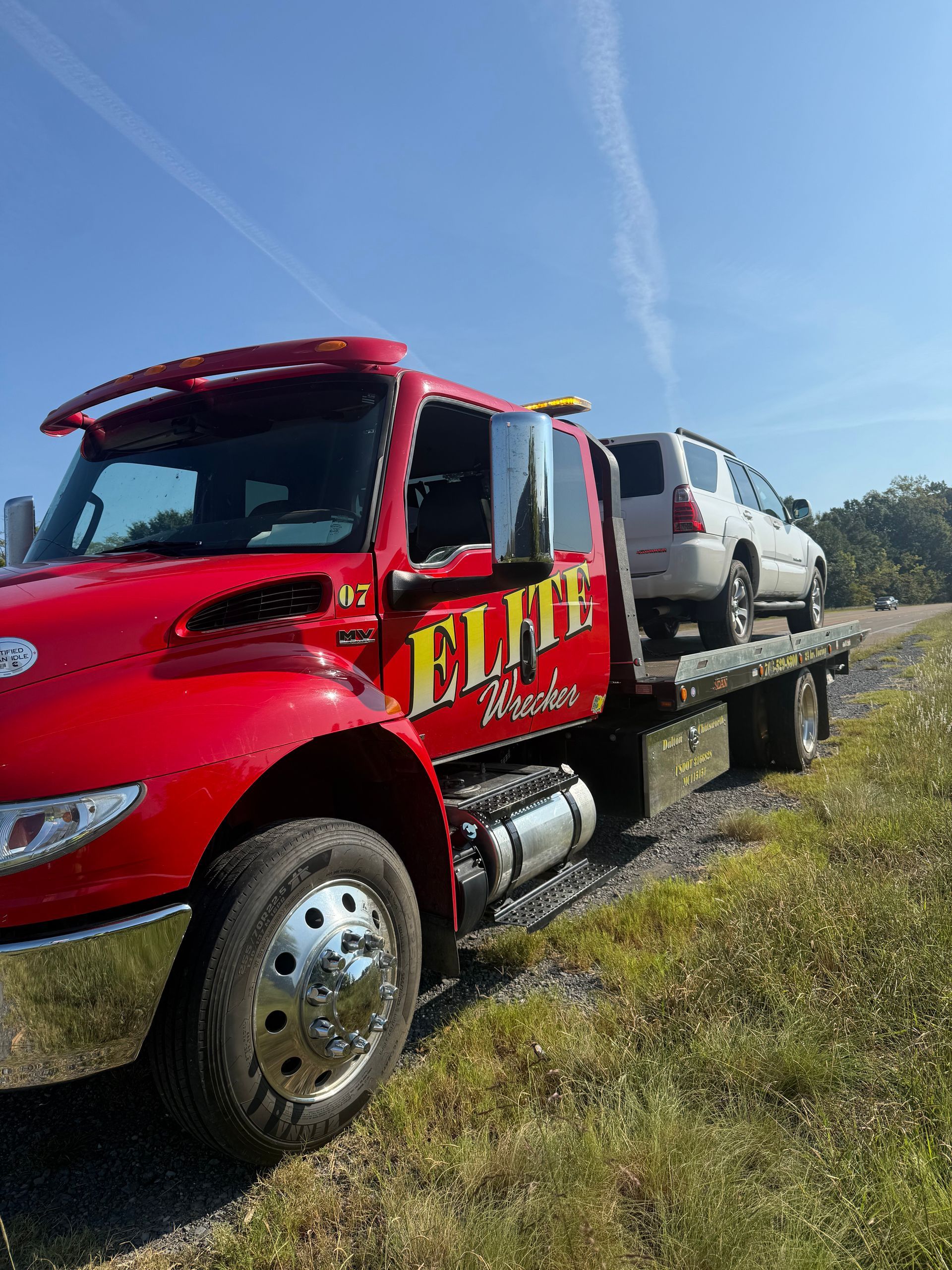 Red tow truck carrying a silver SUV on a flatbed beside a grassy roadside under a blue sky