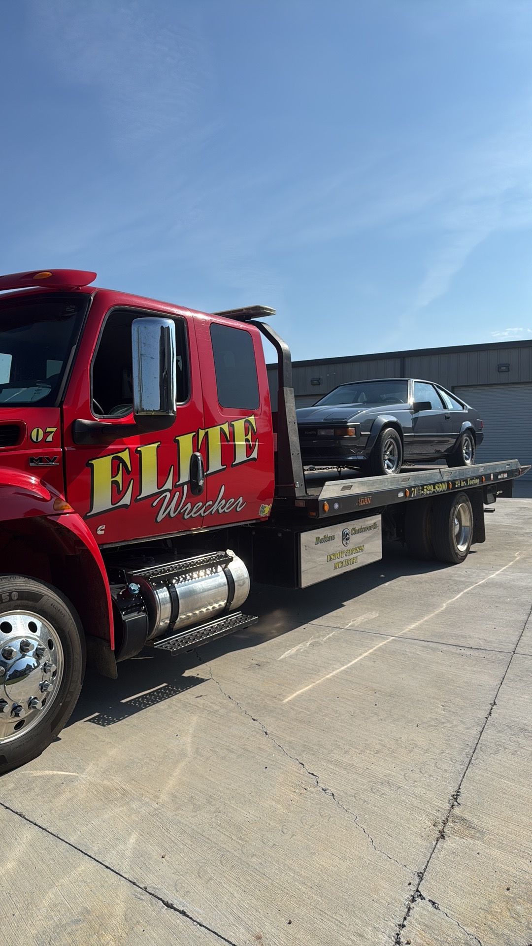 Red ELITE tow truck with a black car on its flatbed in a parking lot under a blue sky