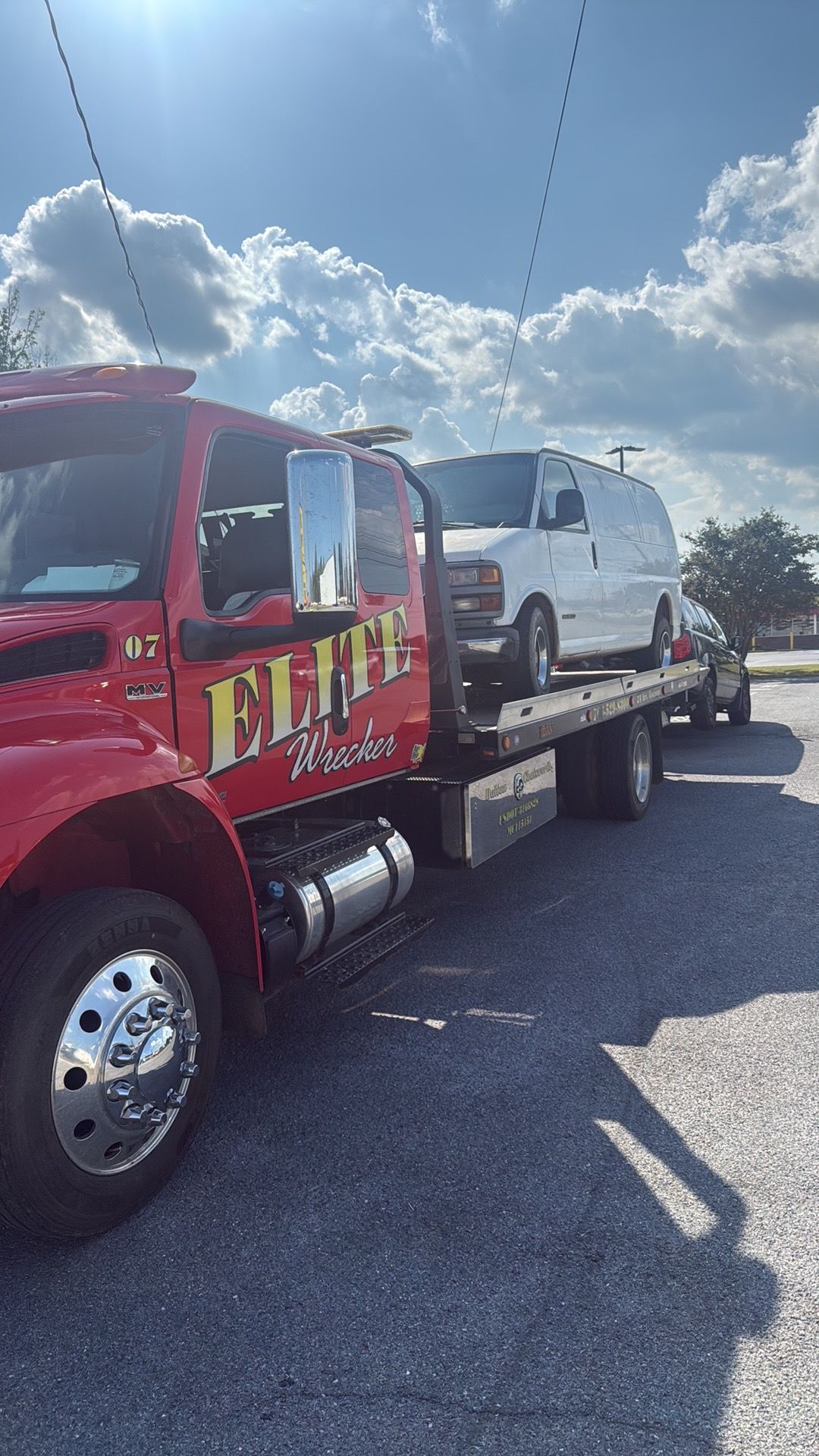Red tow truck carrying a white SUV on a flatbed in a sunny parking lot