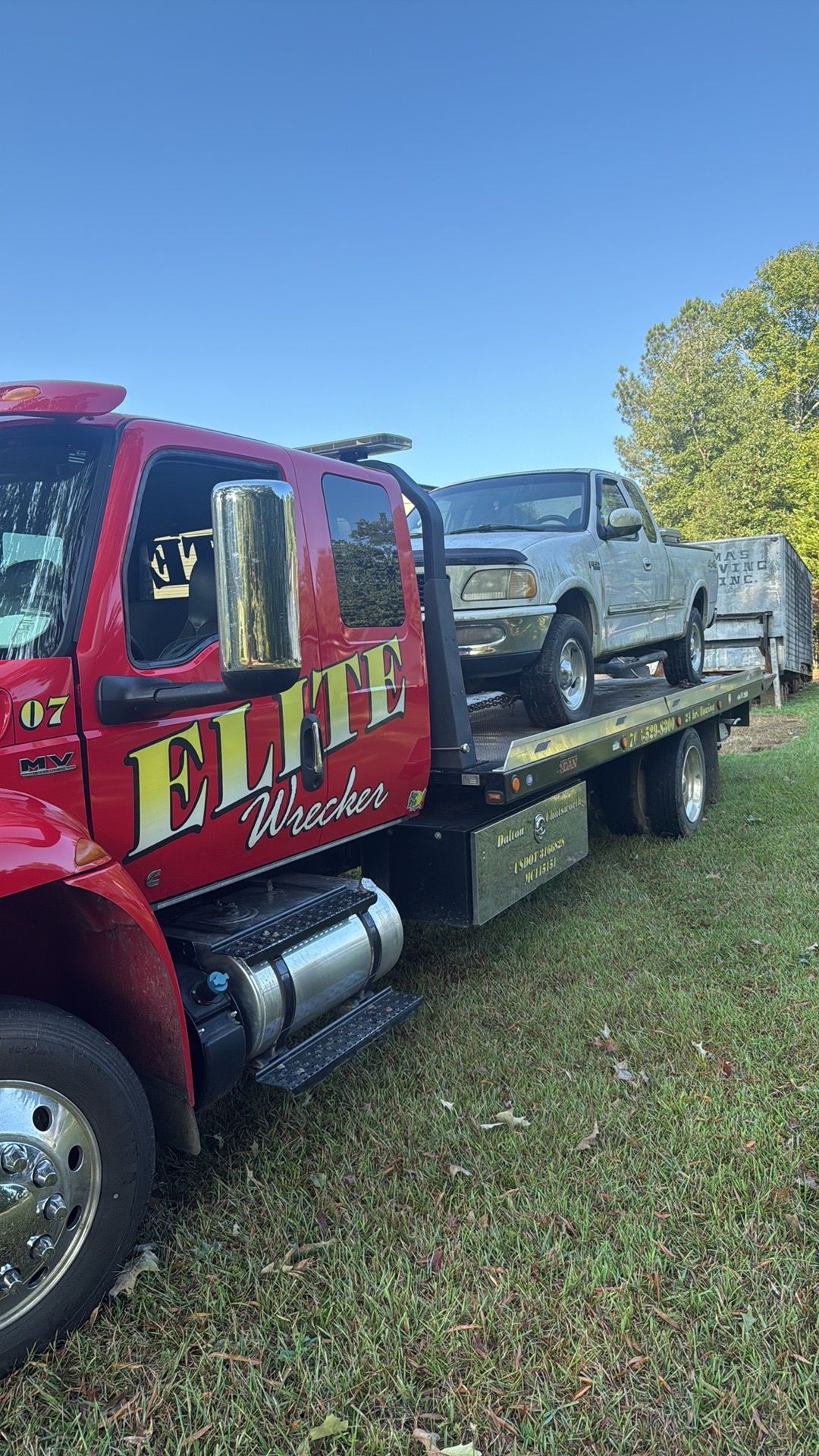 Red tow truck hauling a silver pickup and trailer on grass under a blue sky