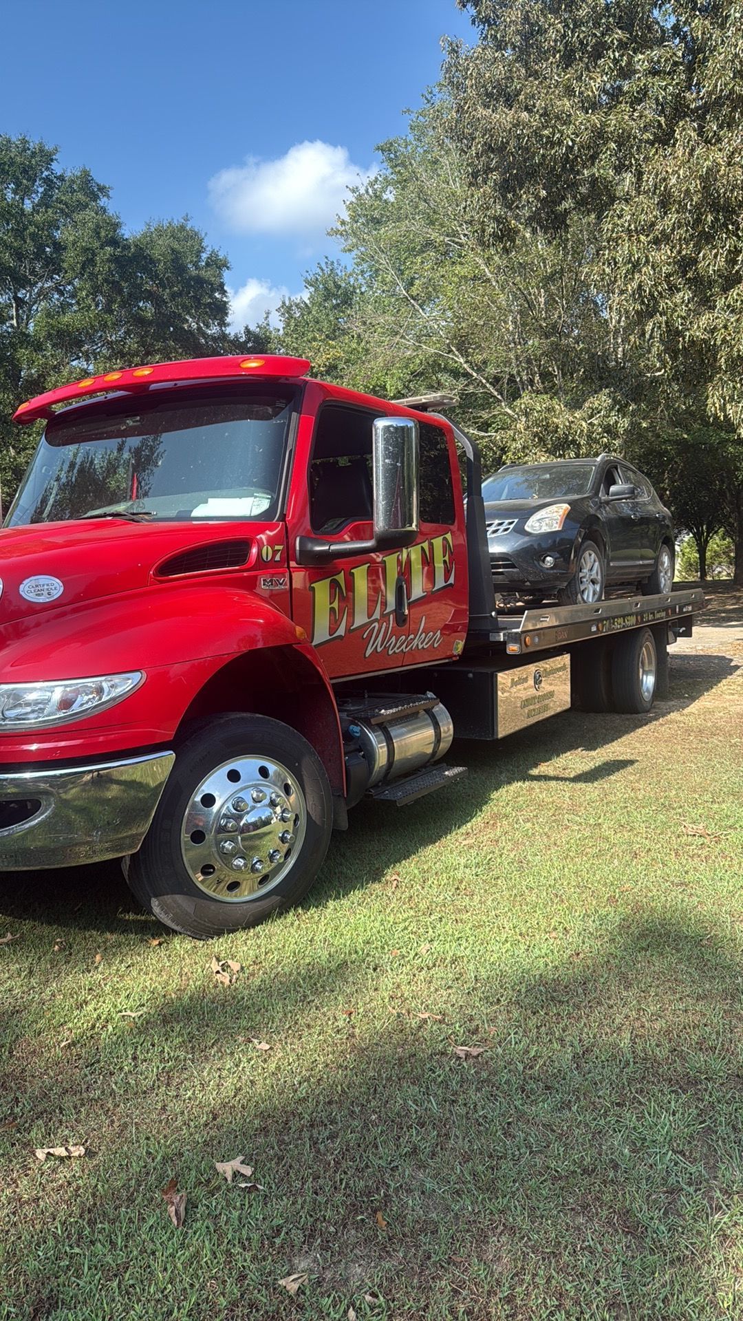 Red flatbed tow truck carrying a black car on a grassy yard with trees in the background