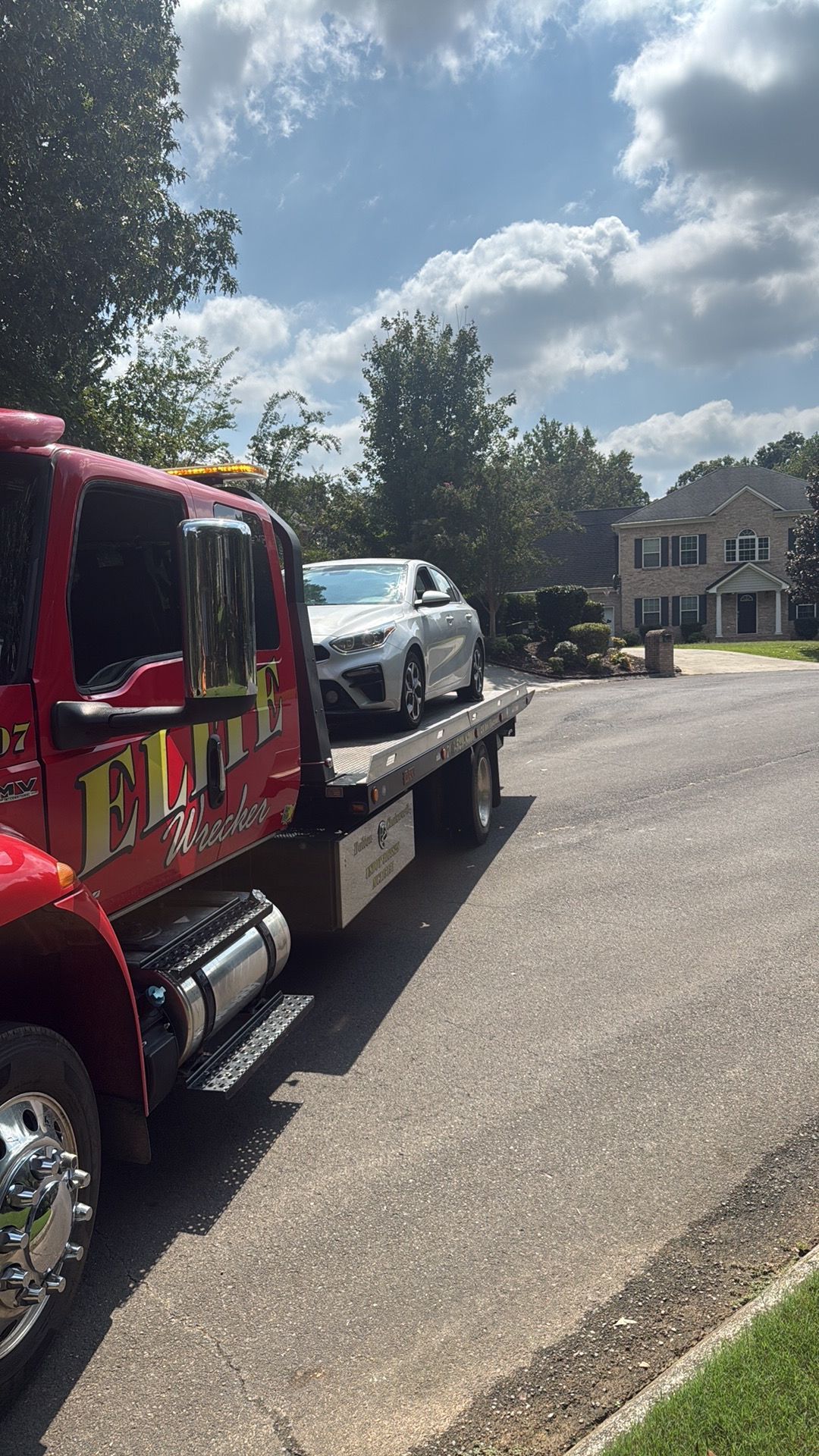 Red tow truck carrying a silver car on a suburban street under a bright cloudy sky