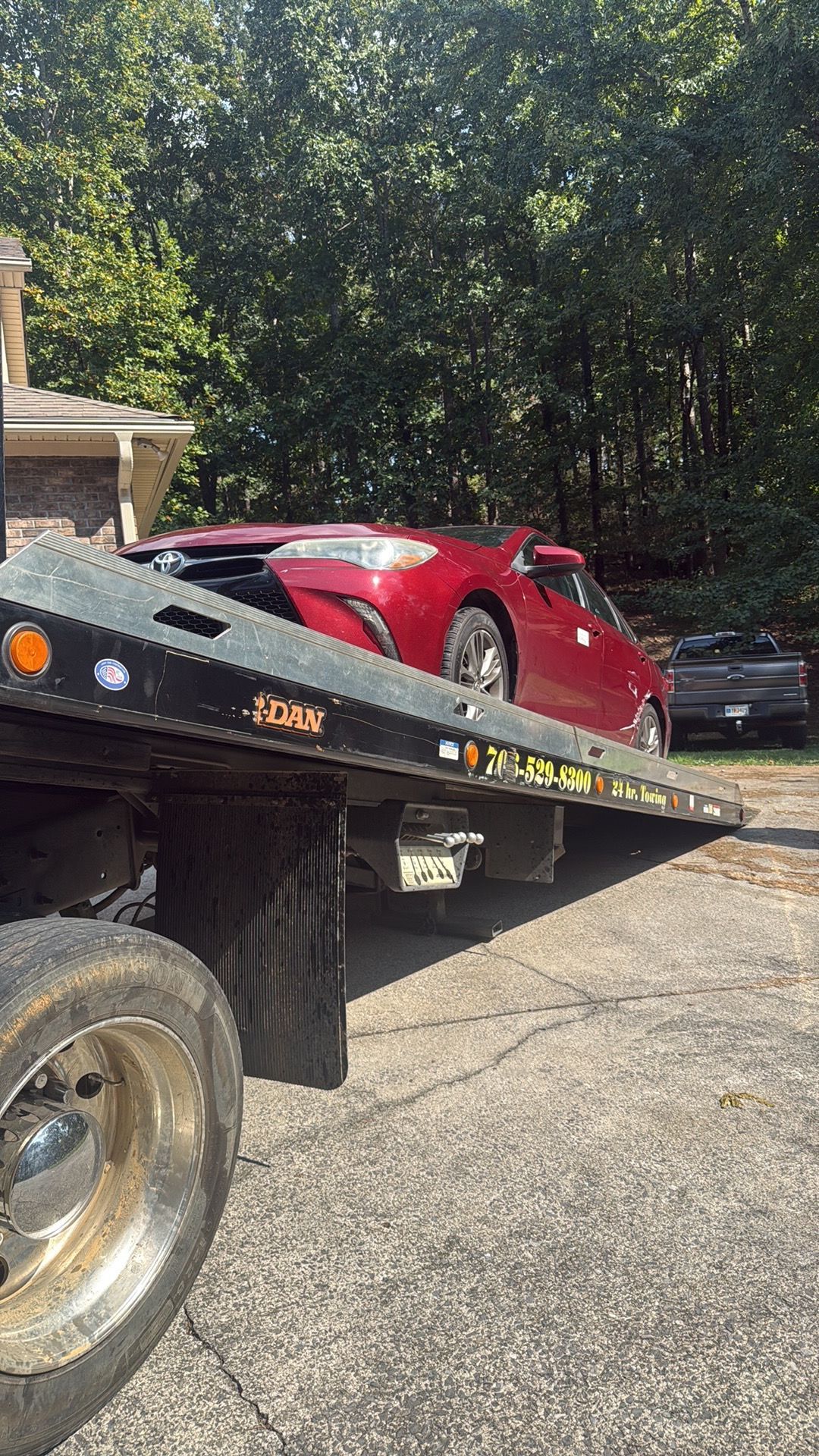 Red car on a flatbed tow truck in a wooded driveway area.