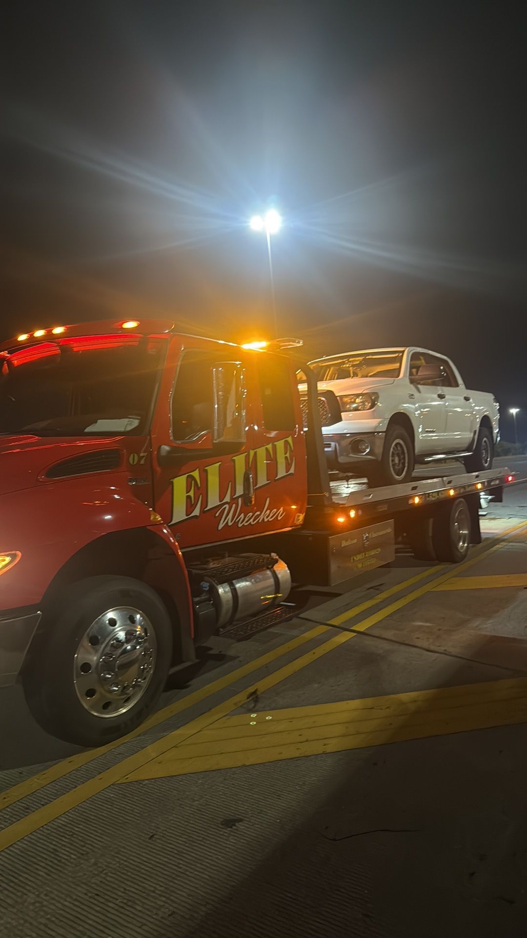 Tow truck hauling a white pickup at night on a city street, with flashing amber and red lights.