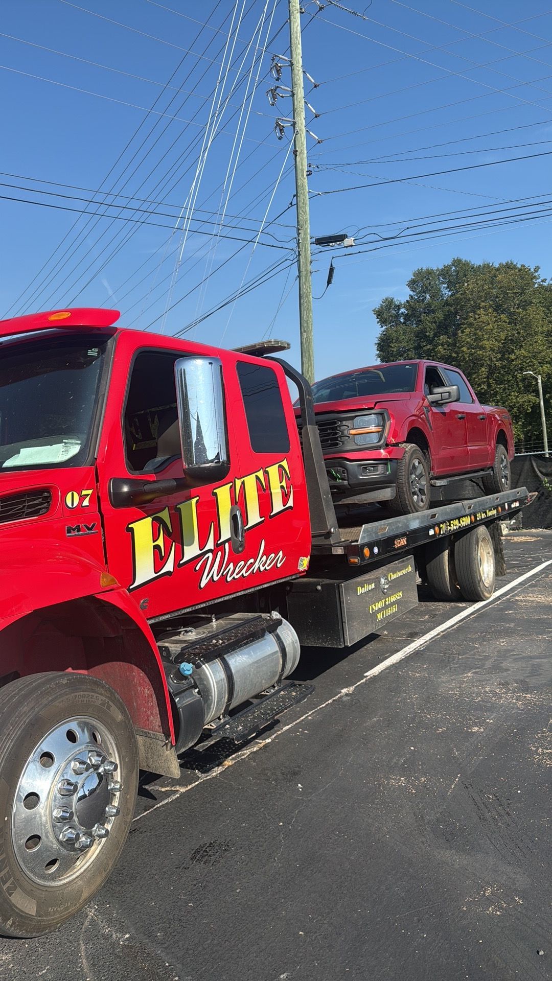 Red tow truck carrying a red pickup on a flatbed, parked by a road under a clear blue sky.