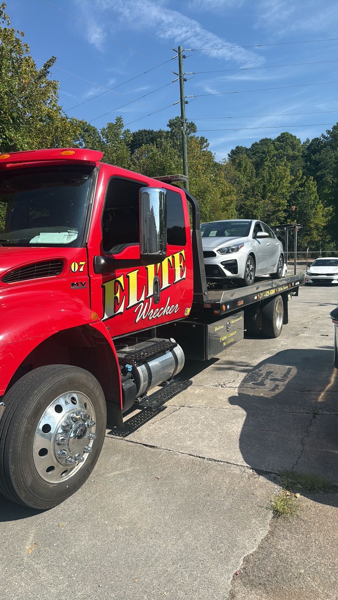 Red tow truck carrying a white car on a flatbed in a sunny outdoor lot