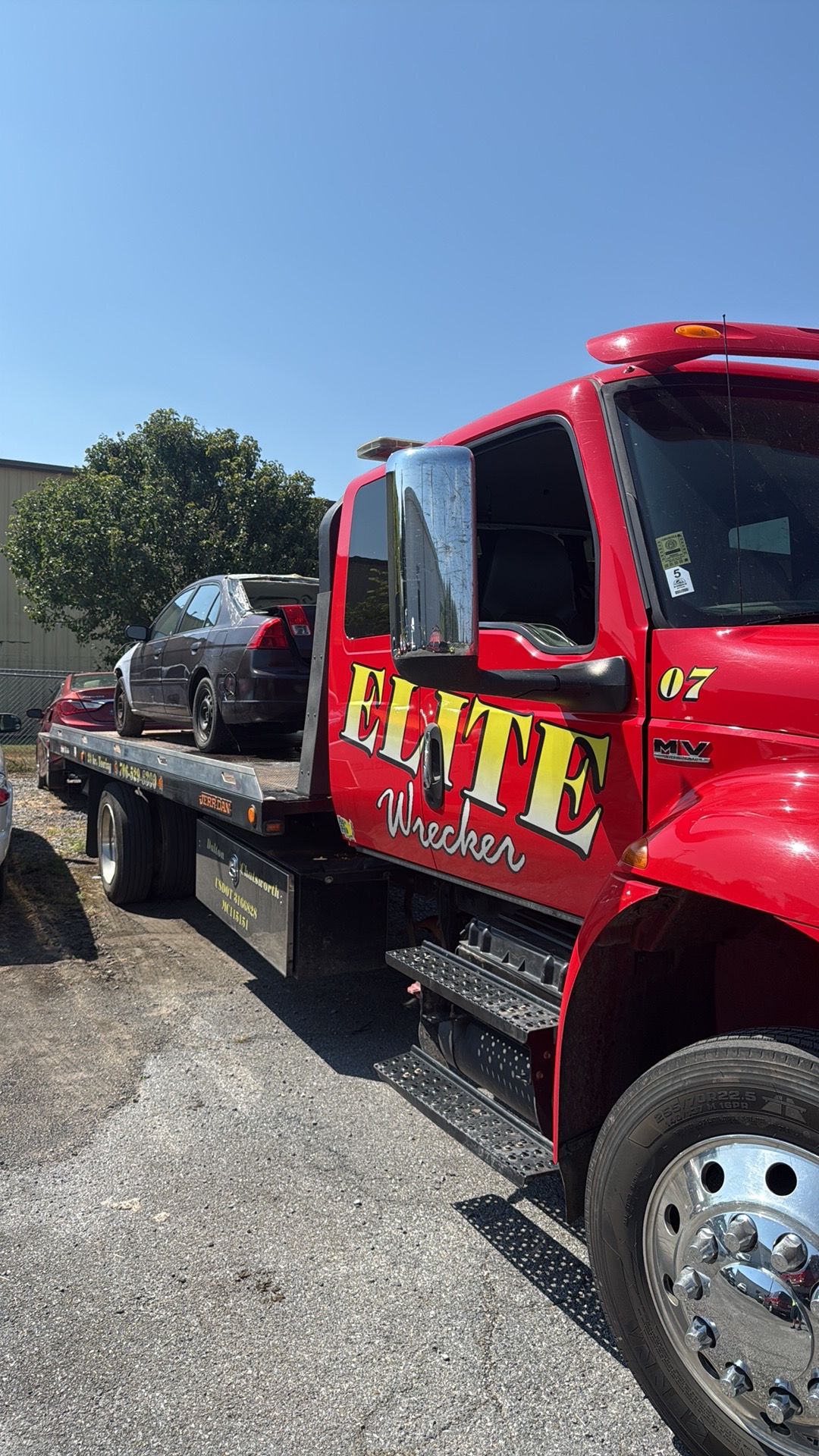Red tow truck loading a gray car on a flatbed in a sunny parking lot.