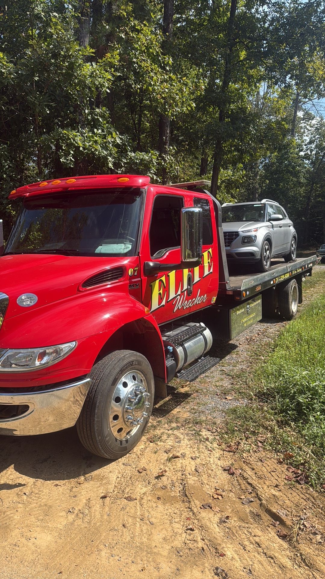Red tow truck carrying a silver SUV on a dirt road beside trees