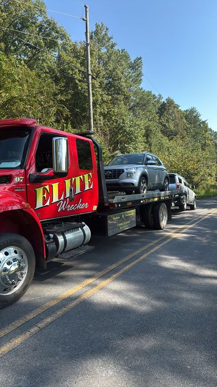 Red tow truck hauling a black car on a tree-lined road under a clear blue sky