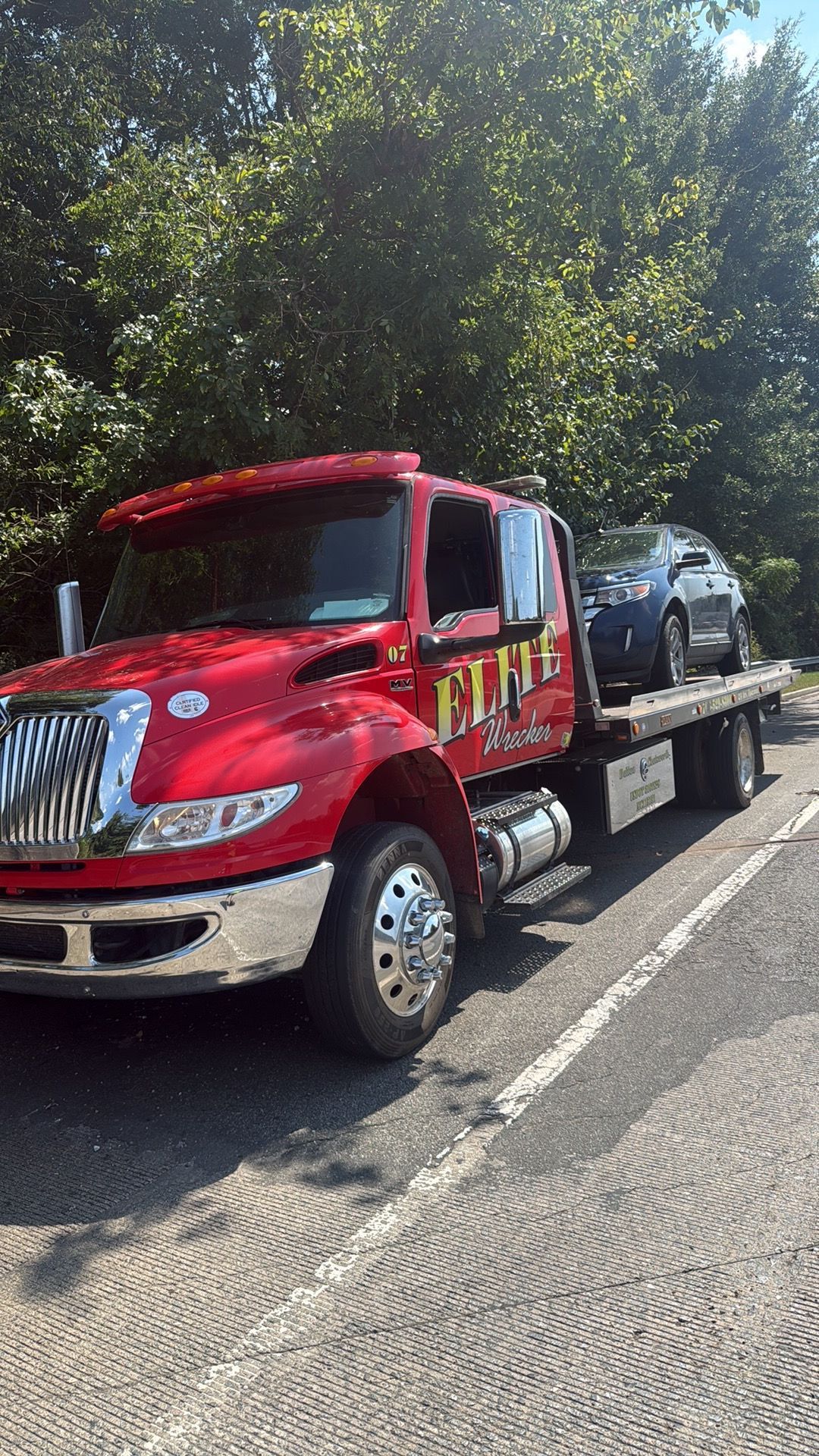 Red tow truck hauling a black car on a flatbed beside a tree-lined road.