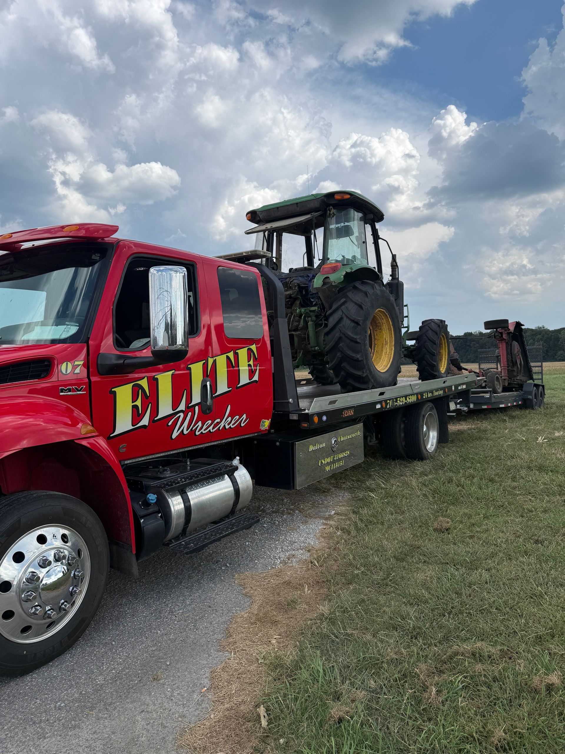 Red tow truck hauling a tractor on a flatbed trailer beside a road under a cloudy sky