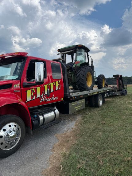 Red tow truck hauling a tractor on a flatbed along a rural roadside under a cloudy sky
