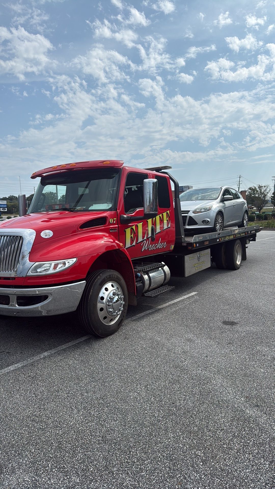 Red tow truck hauling a silver car on a roadside under a partly cloudy sky