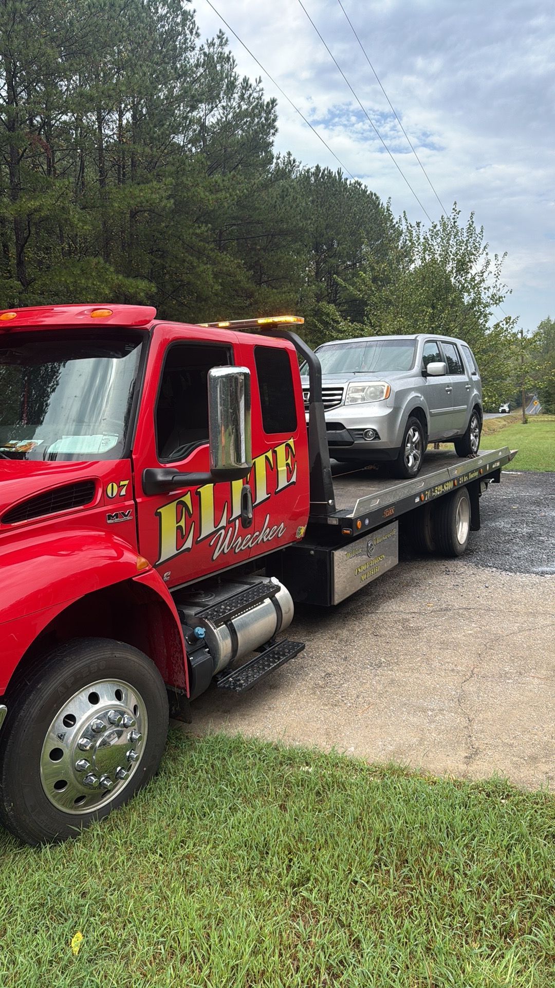 Red tow truck hauling a silver SUV on a flatbed by a roadside with trees and grass nearby
