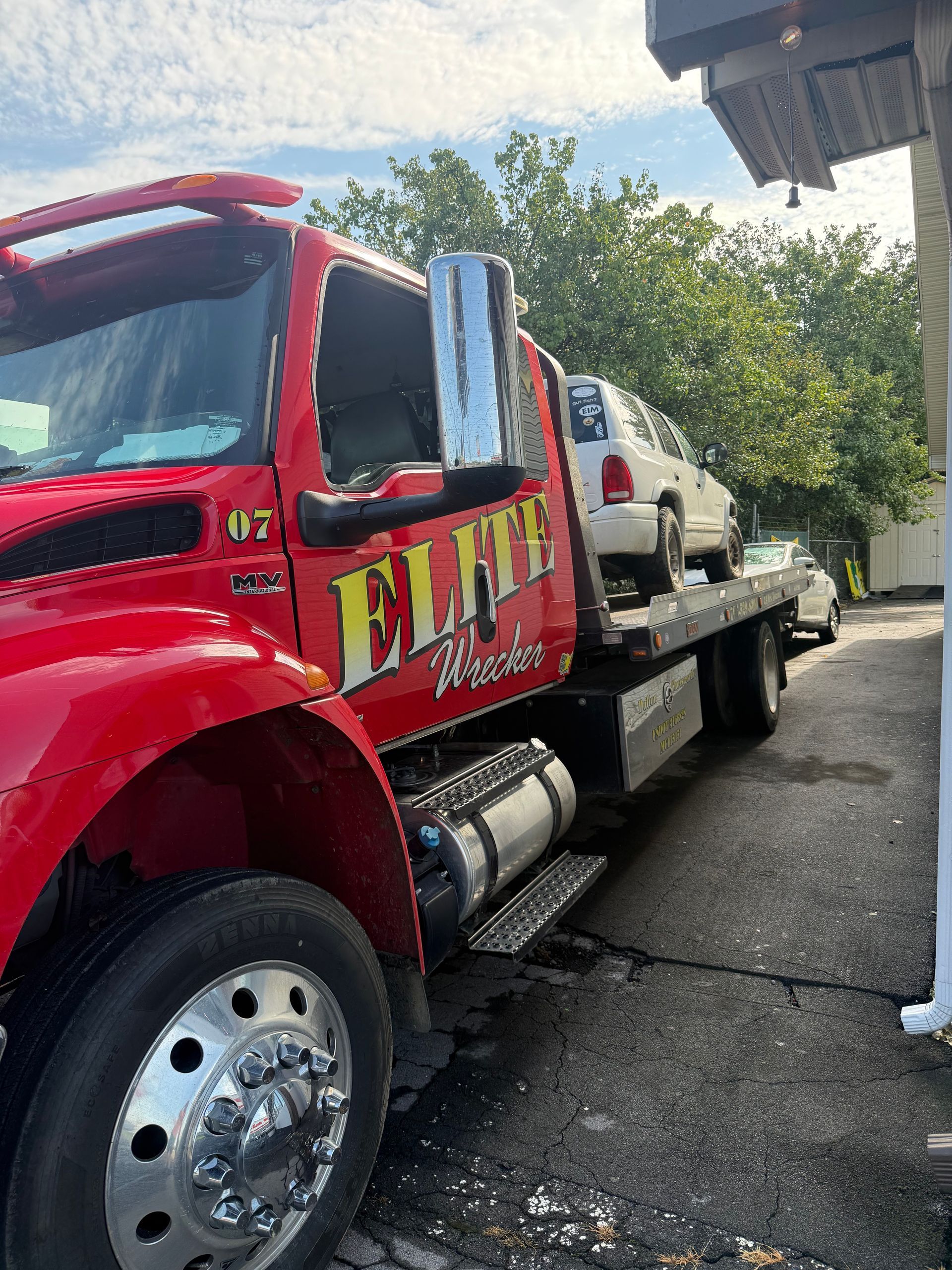 Red tow truck with large “ELITE” lettering carrying a white car on a flatbed by a building and trees.
