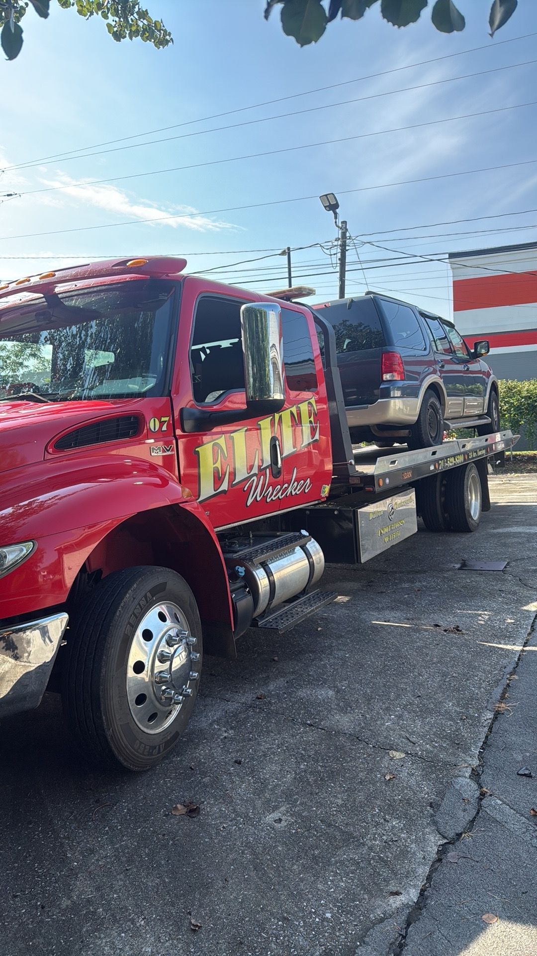 Red tow truck hauling a black SUV on a flatbed in a parking lot under a blue sky.