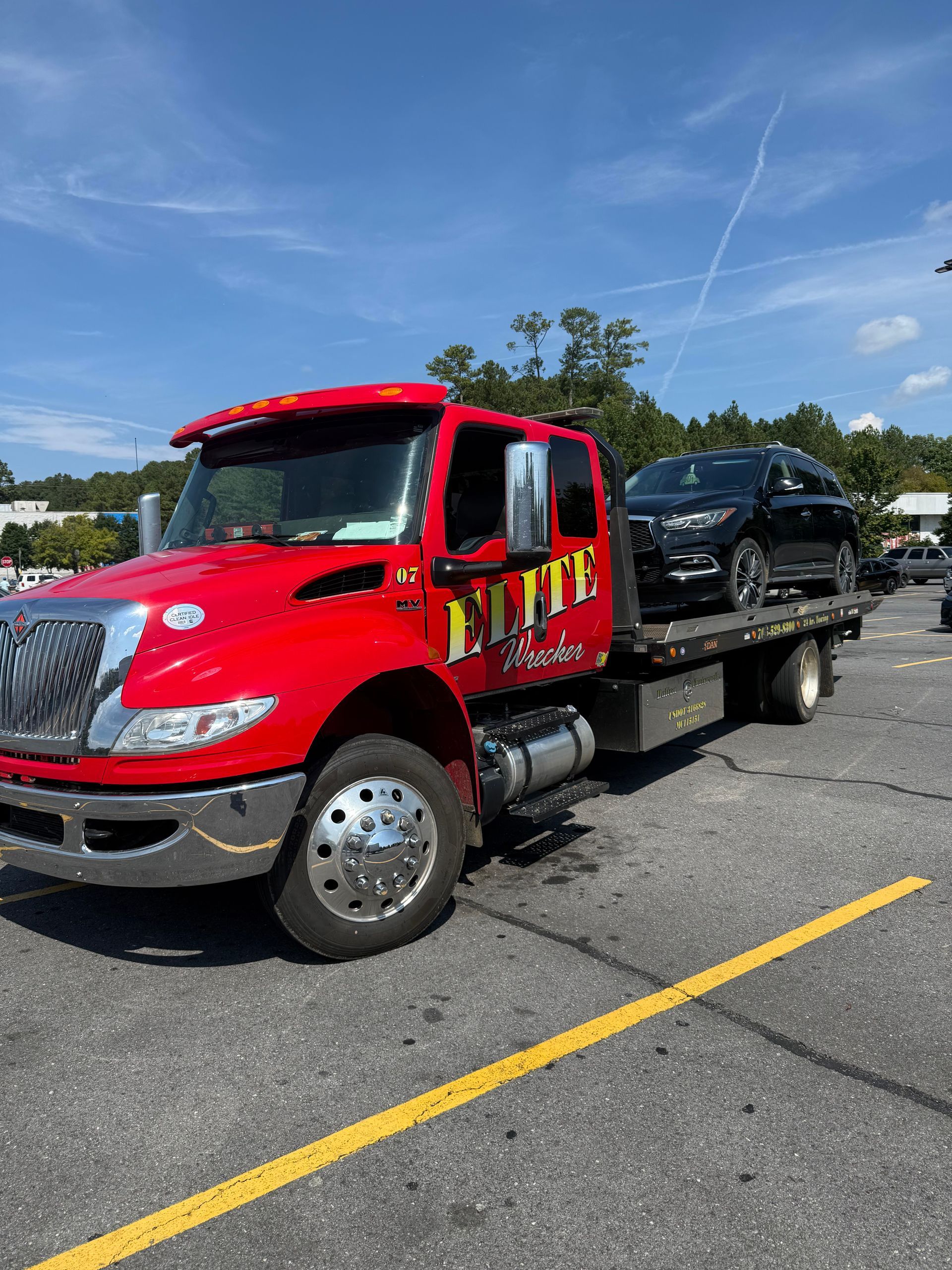 Red tow truck carrying a black SUV on a flatbed in a parking lot under a blue sky